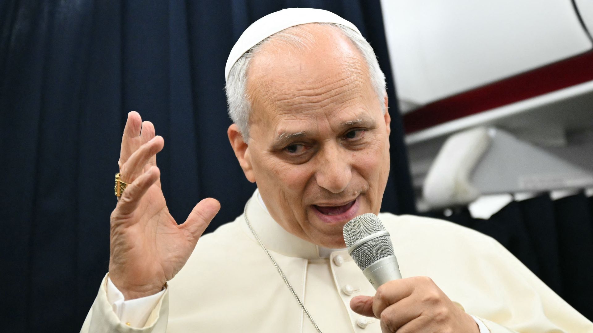 Pope Leo speaks into a microphone while wearing white robes on a plane.