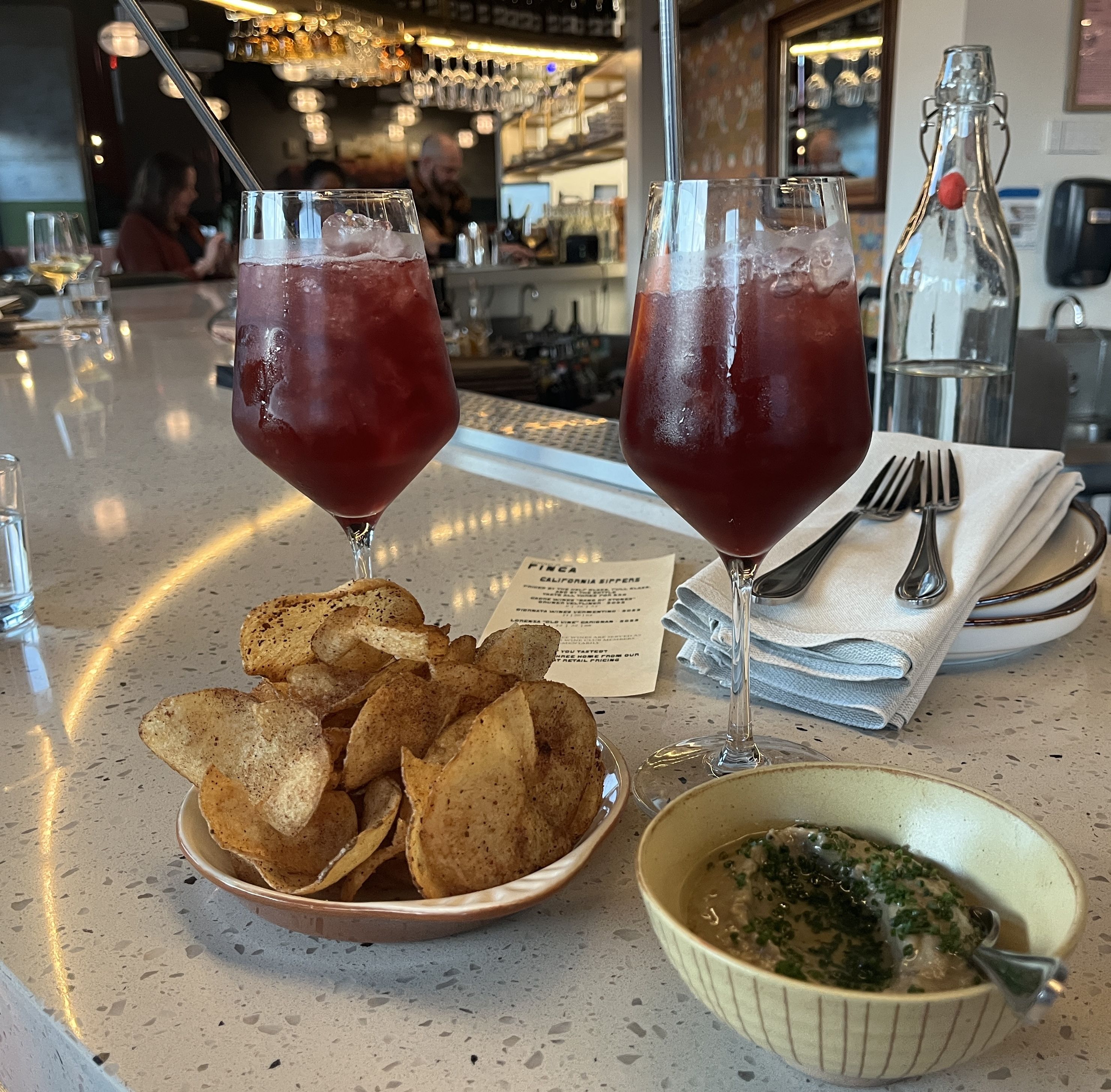 Two tall glasses of red sangria with straws on a bar counter, next to a bowl of seasoned potato chips and a bowl of dip garnished with herbs, with a bar and people in the background.
