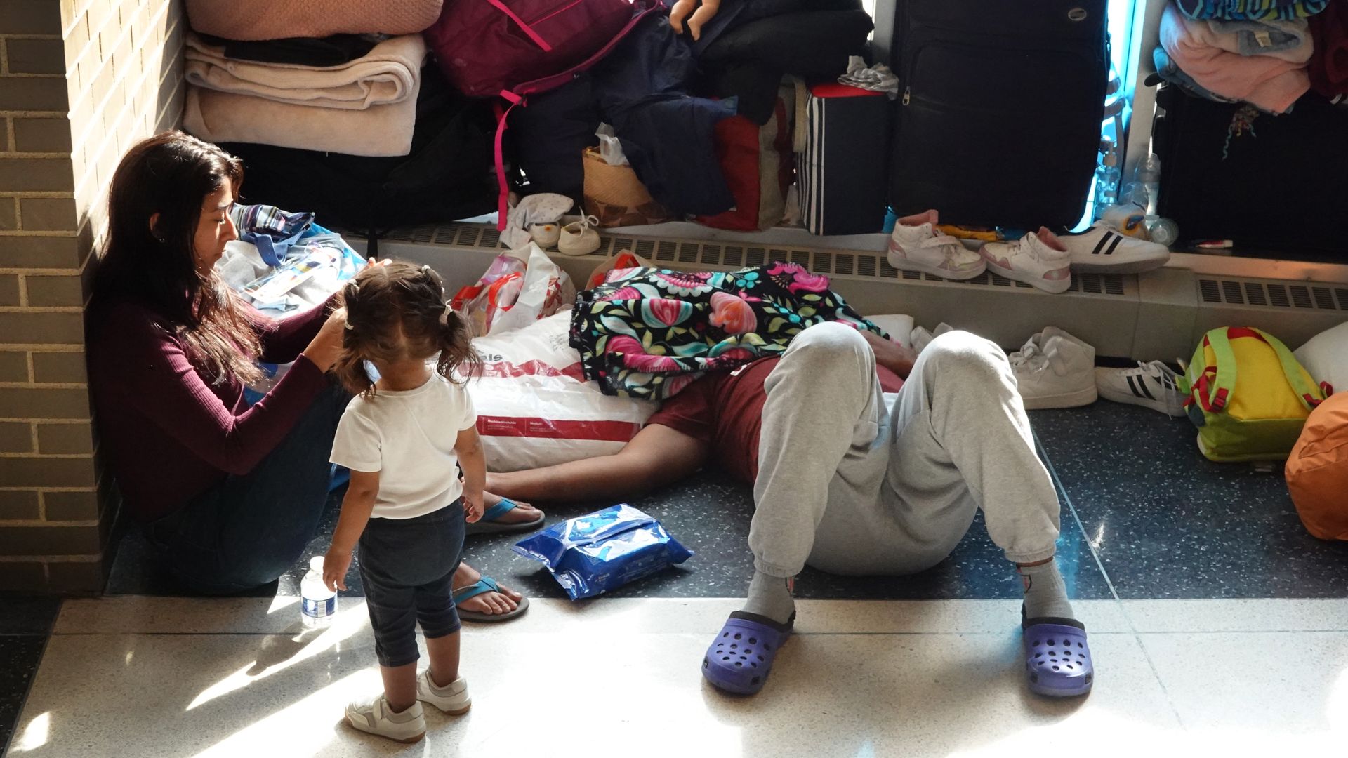 Woman sits on floor, back of a toddler with pigtails, man in sweatpants lies on floor with face covered.