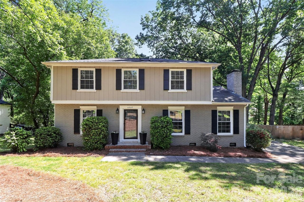 Two-story suburban home at 6527 with beige upper siding and gray brick lower, black shutters, central front door with brick steps, and tall green trees surrounding the yard.