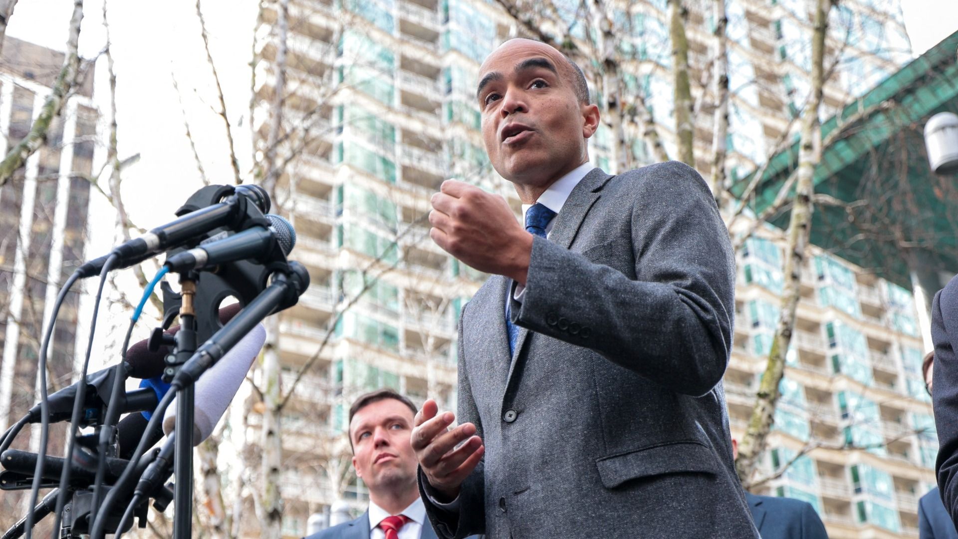 Washington Attorney General Nick Brown speaks in front of a downtown apartment building and a courthouse, with Noah Purcell in the background. Brown is talking into a collection of microphones at a press conference while gesturing with his hands.