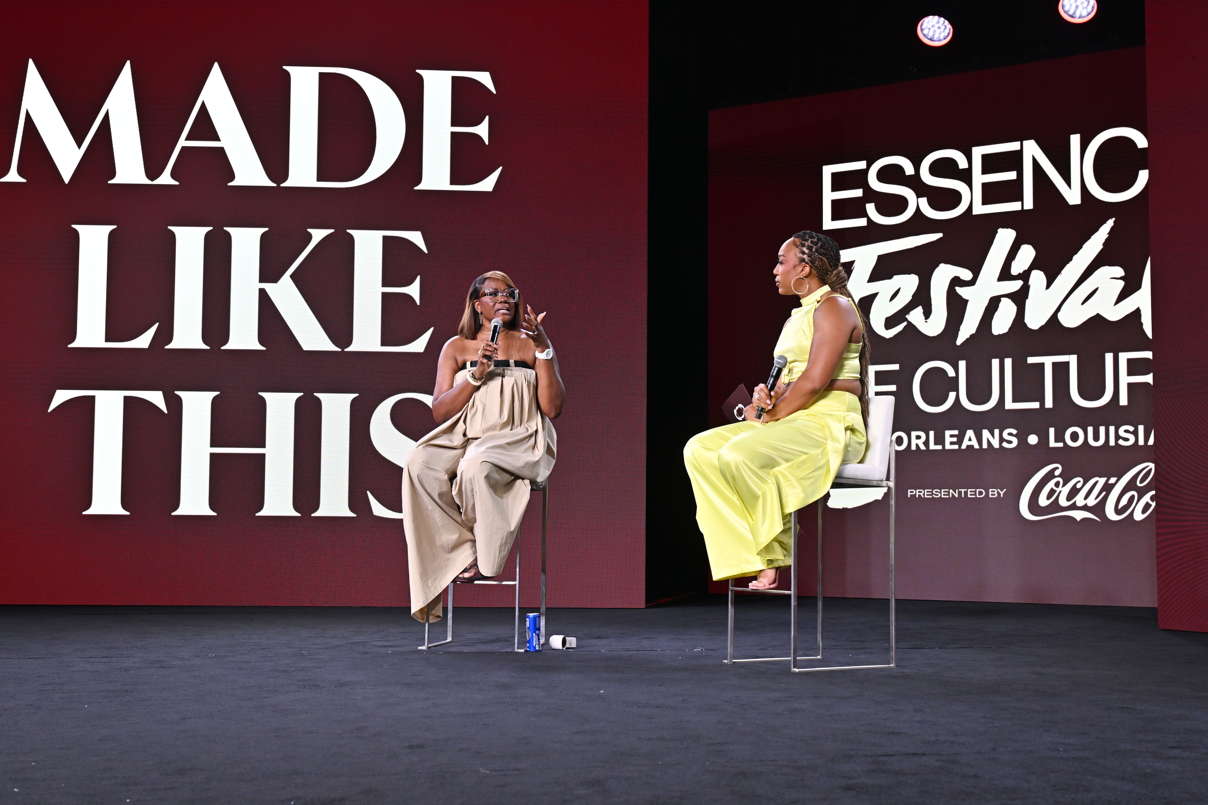 Two women seated on high stools speaking at the Essence Festival of Culture event in New Orleans. One wears a beige strapless dress, the other a yellow sleeveless outfit.