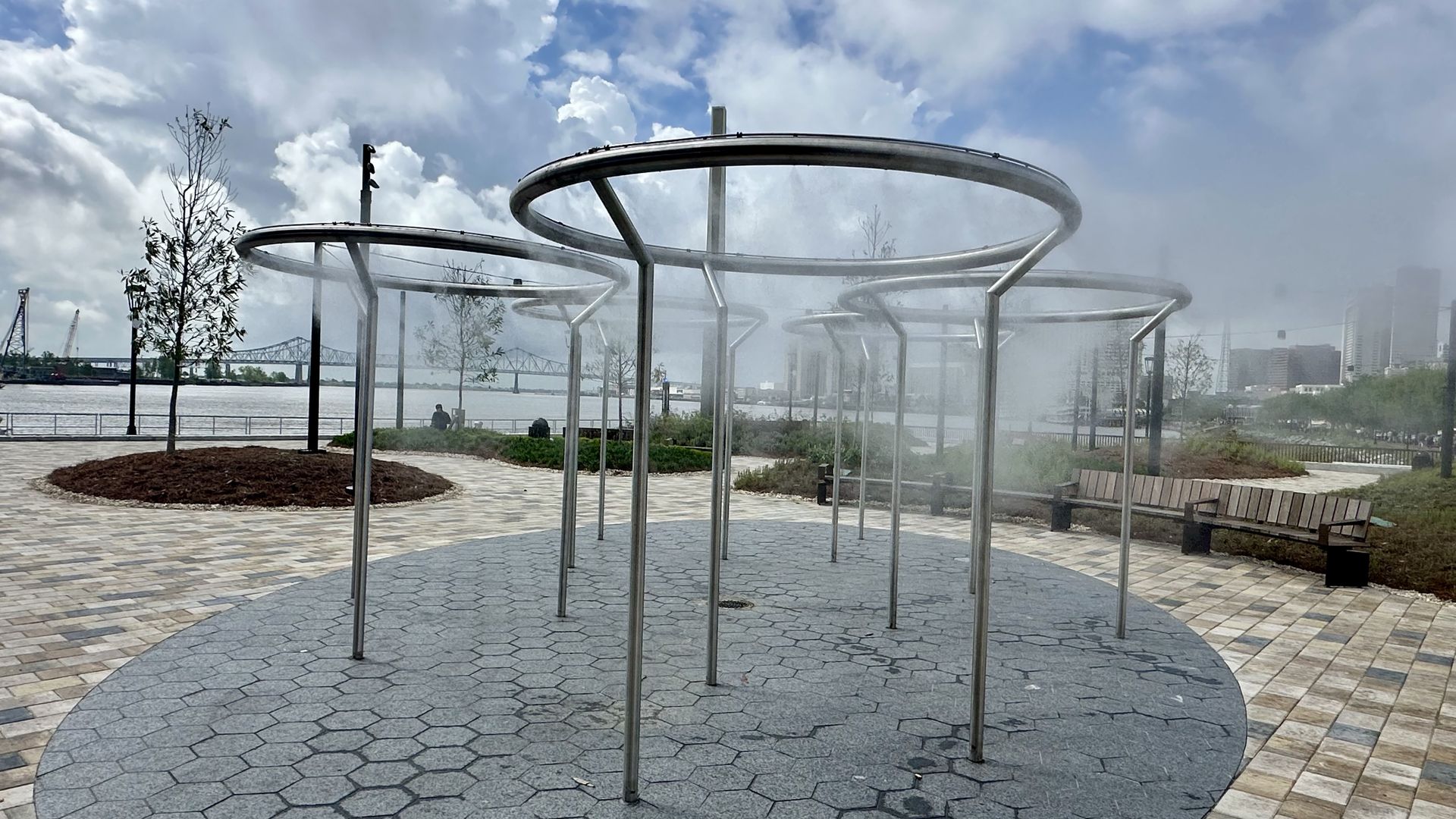 Riverside park with a metallic sculpture of large circular rings on posts; mist streams through rings above a hexagonal-tiled plaza, benches, trees, river, and a distant bridge.