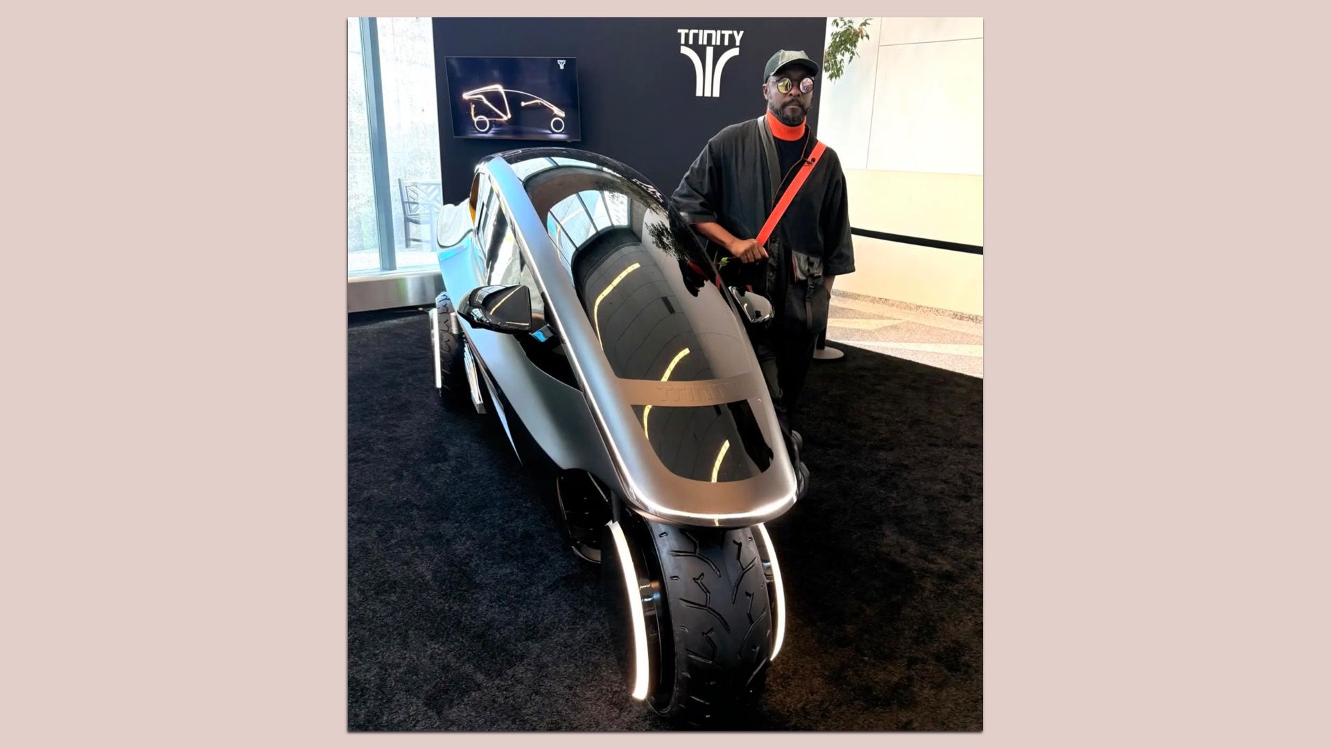 A man in dark attire with an orange crossbody strap stands beside a silver futuristic motorcycle with a curved glass canopy at a Trinity exhibit, with a display screen in the background.