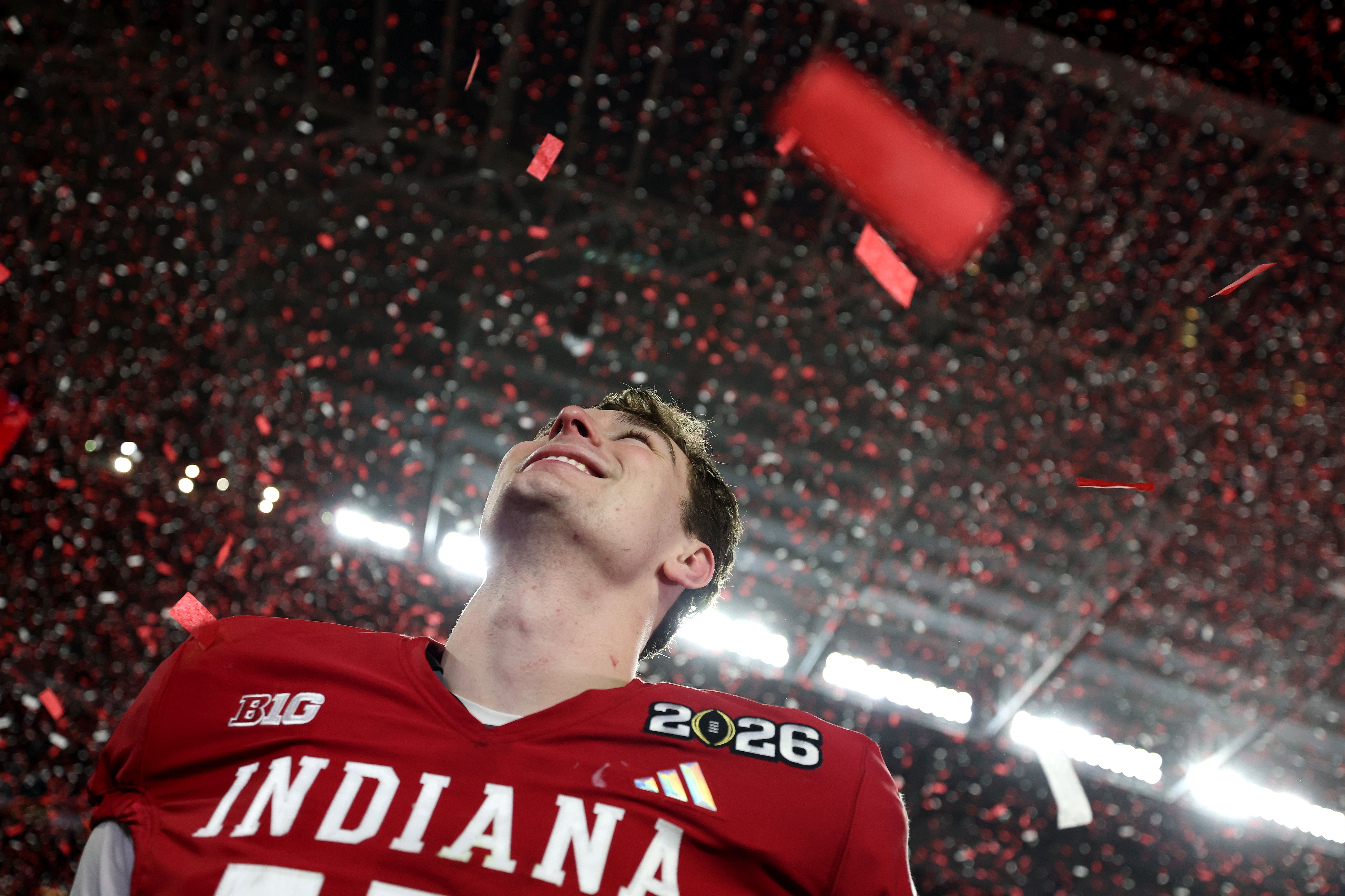 A joyful Indiana football player wearing a red jersey with "2026" patch looks upward amidst falling red confetti under bright stadium lights.