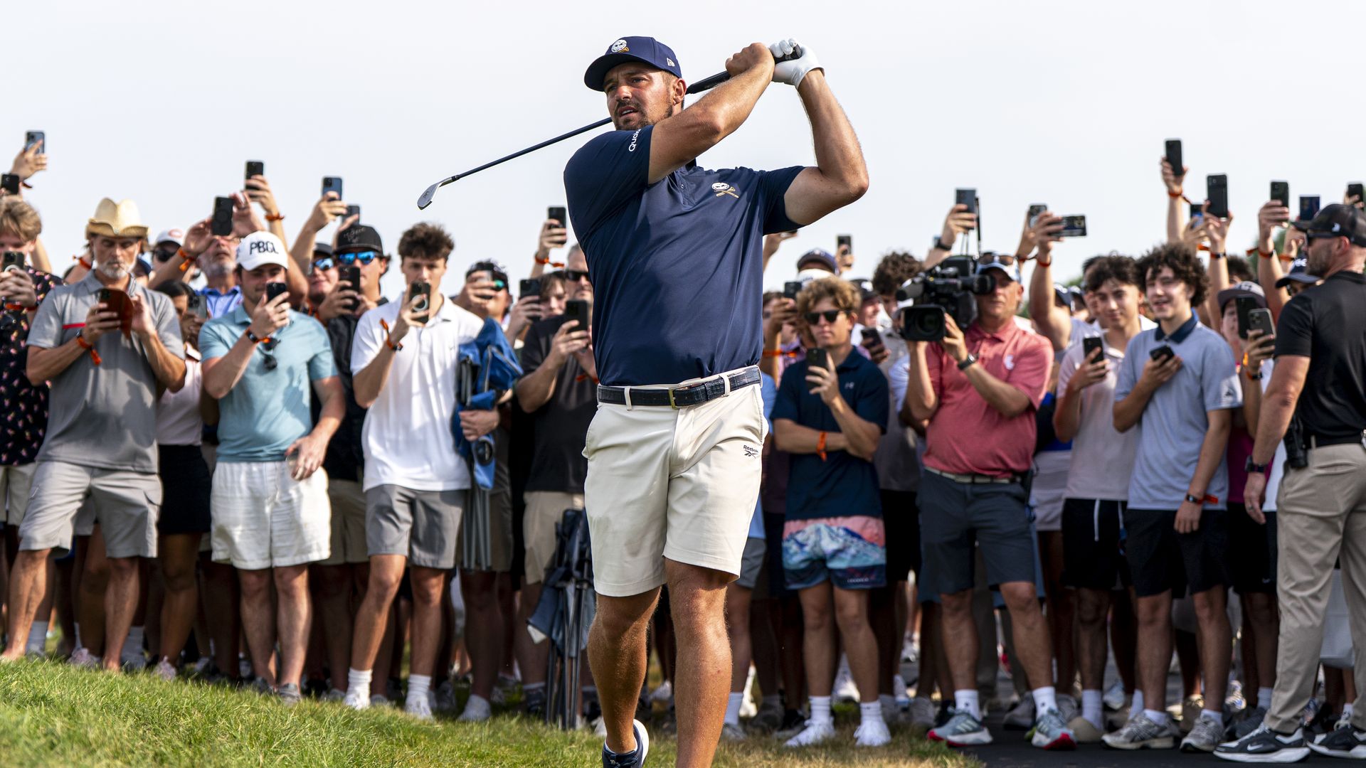 Golfer in navy shirt and white shorts finishing a swing on a green course, surrounded by a crowd taking photos and videos with smartphones.