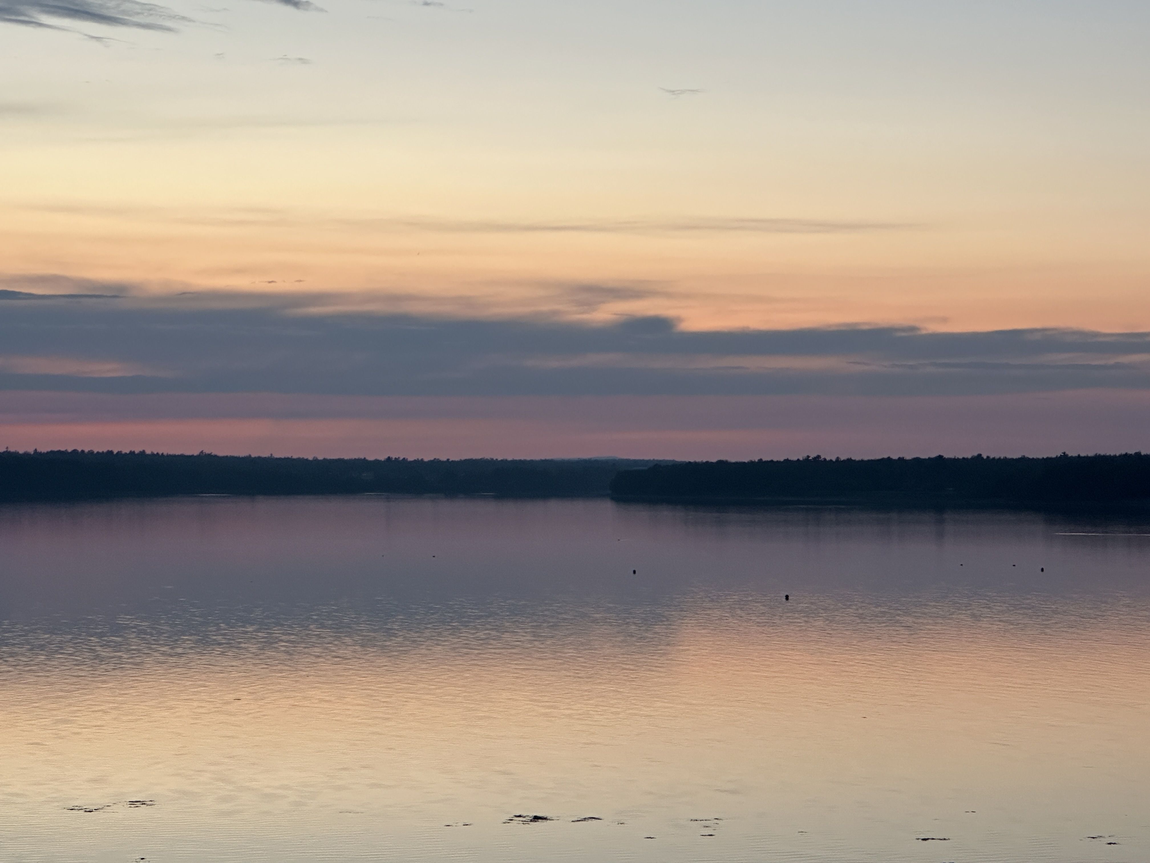 Calm lake reflecting a sunset sky with soft orange, pink, and purple hues, and a dark tree-lined horizon under scattered clouds.