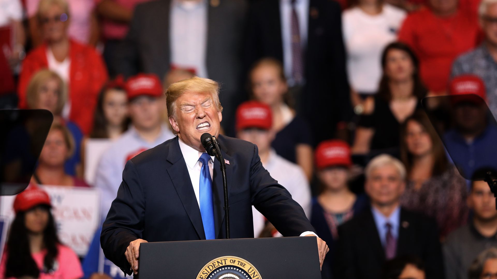 President Trump at a podium with his eyes scrunched shut and his teeth grimacing appearing as if mocking someone or something