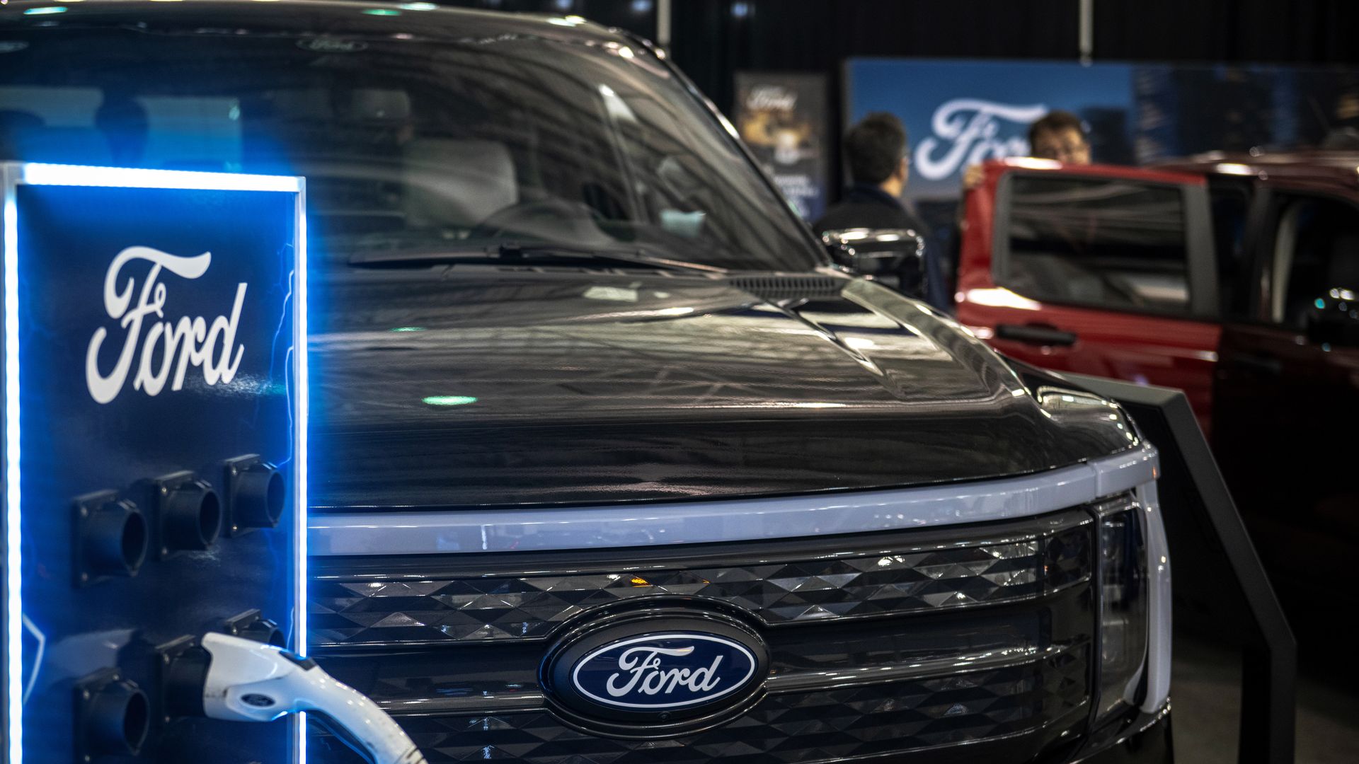 Black Ford electric vehicle being charged at a Ford-branded charging station with blue LED lights, with people and another red vehicle visible in the background.