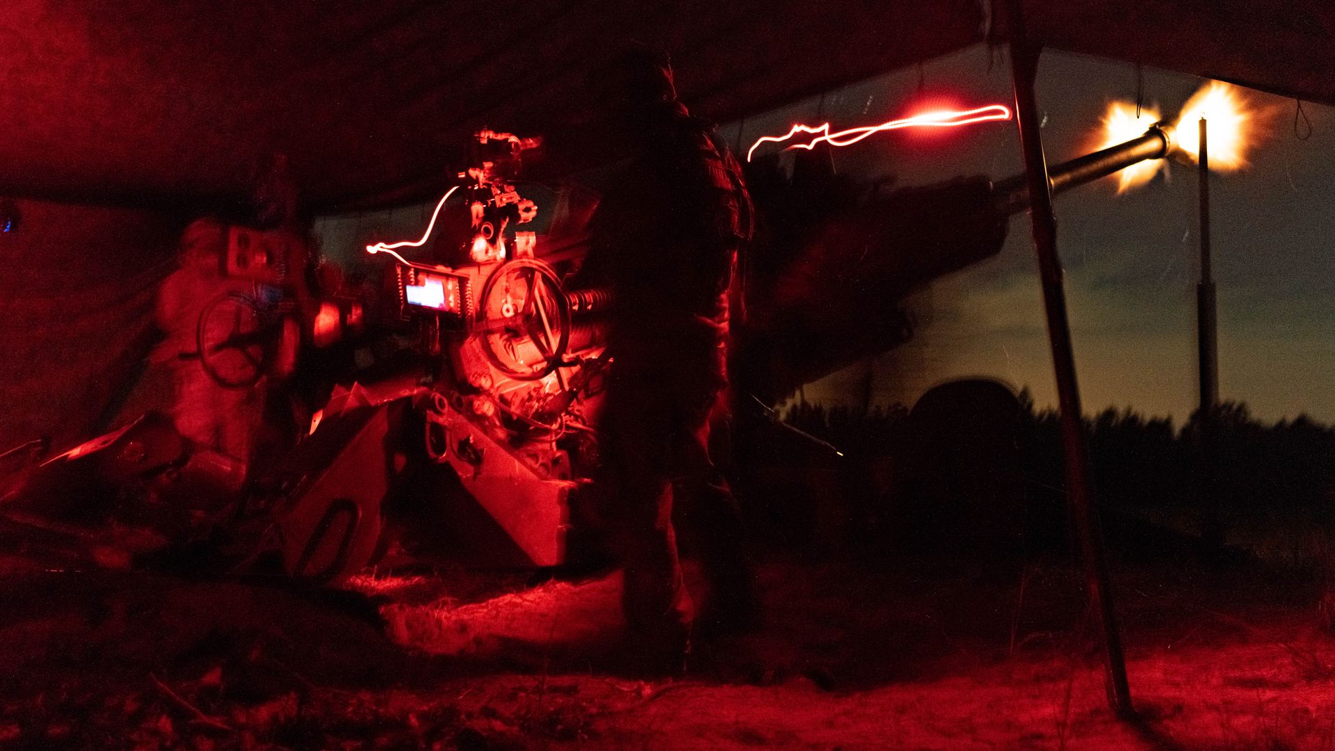 Silhouetted soldier firing a large artillery cannon at night, surrounded by red light and smoke, with a bright muzzle flash and a glowing tracer streak.