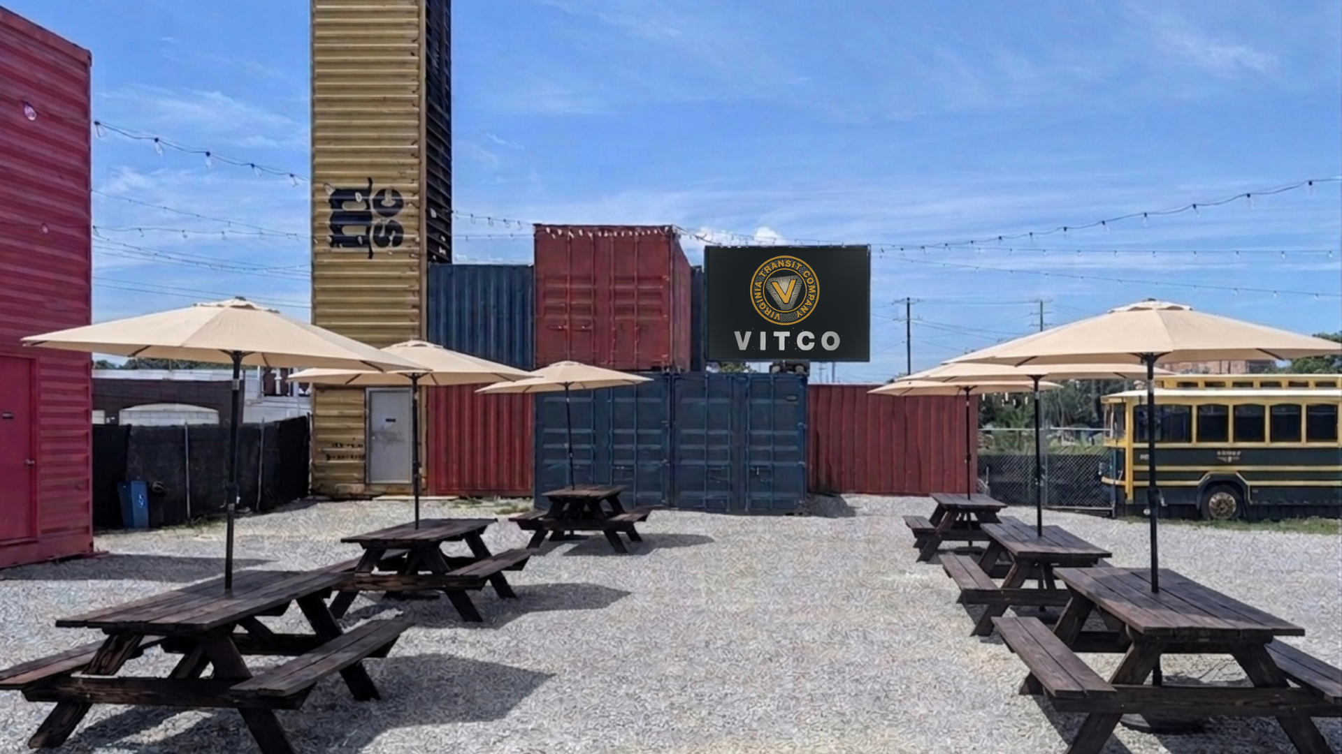 Outdoor yard with stacked shipping containers in red, yellow, and blue; string lights; beige umbrellas over wooden picnic tables; a black sign reading "VITCO" with a circular emblem against clear sky.