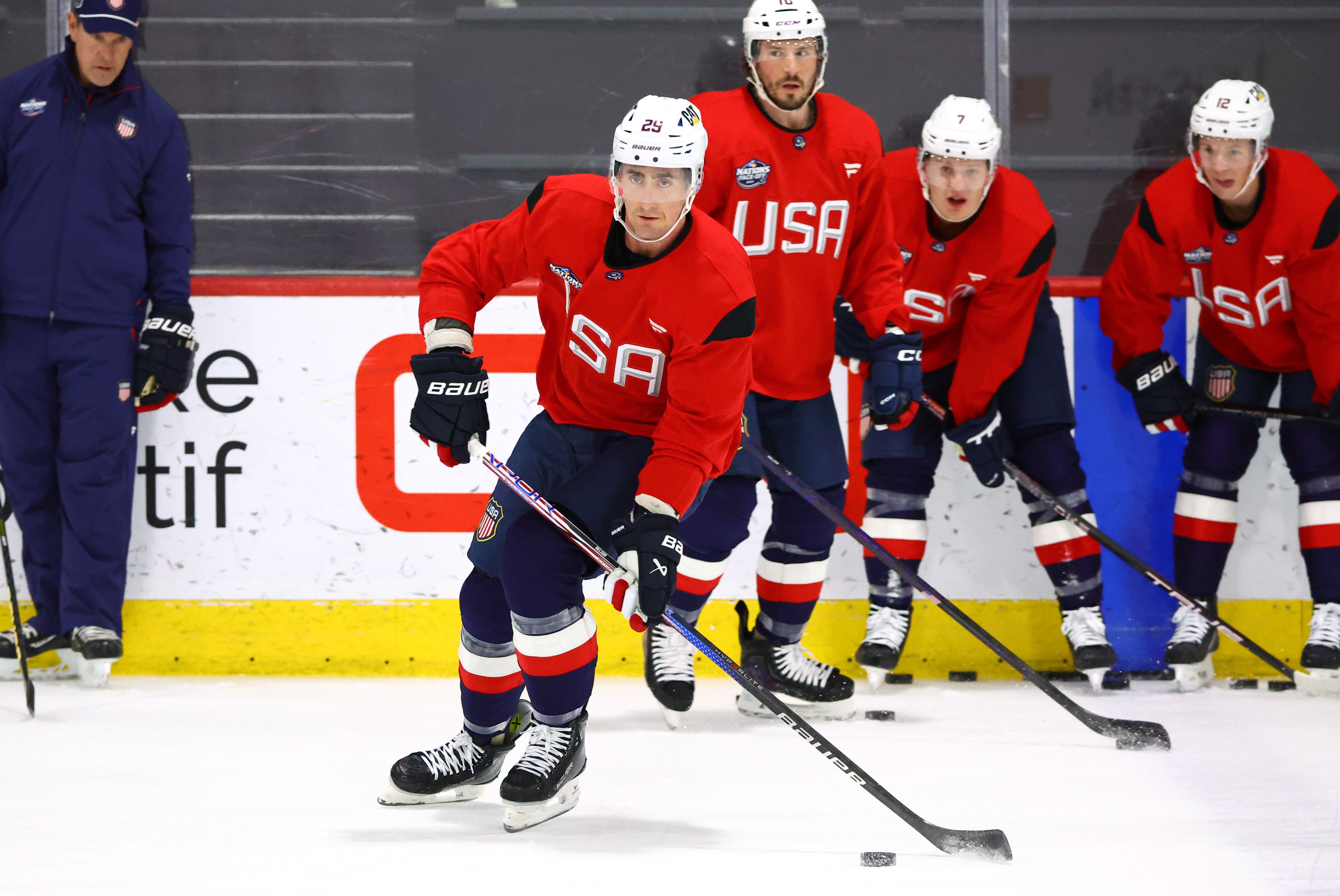 A hockey player in a red jersey skates with the puck during practice as other players look on