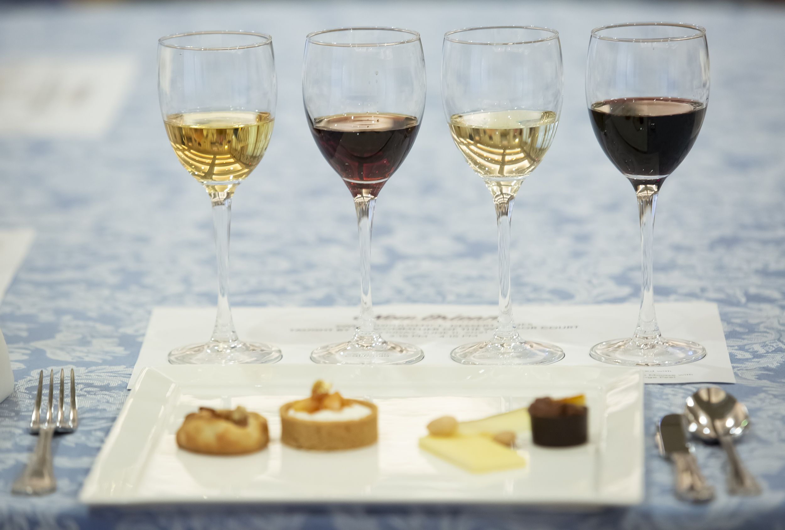 Photo shows wine glasses lined up and filled with different colored wines. A plate of small bites is on the table in front of the wines.