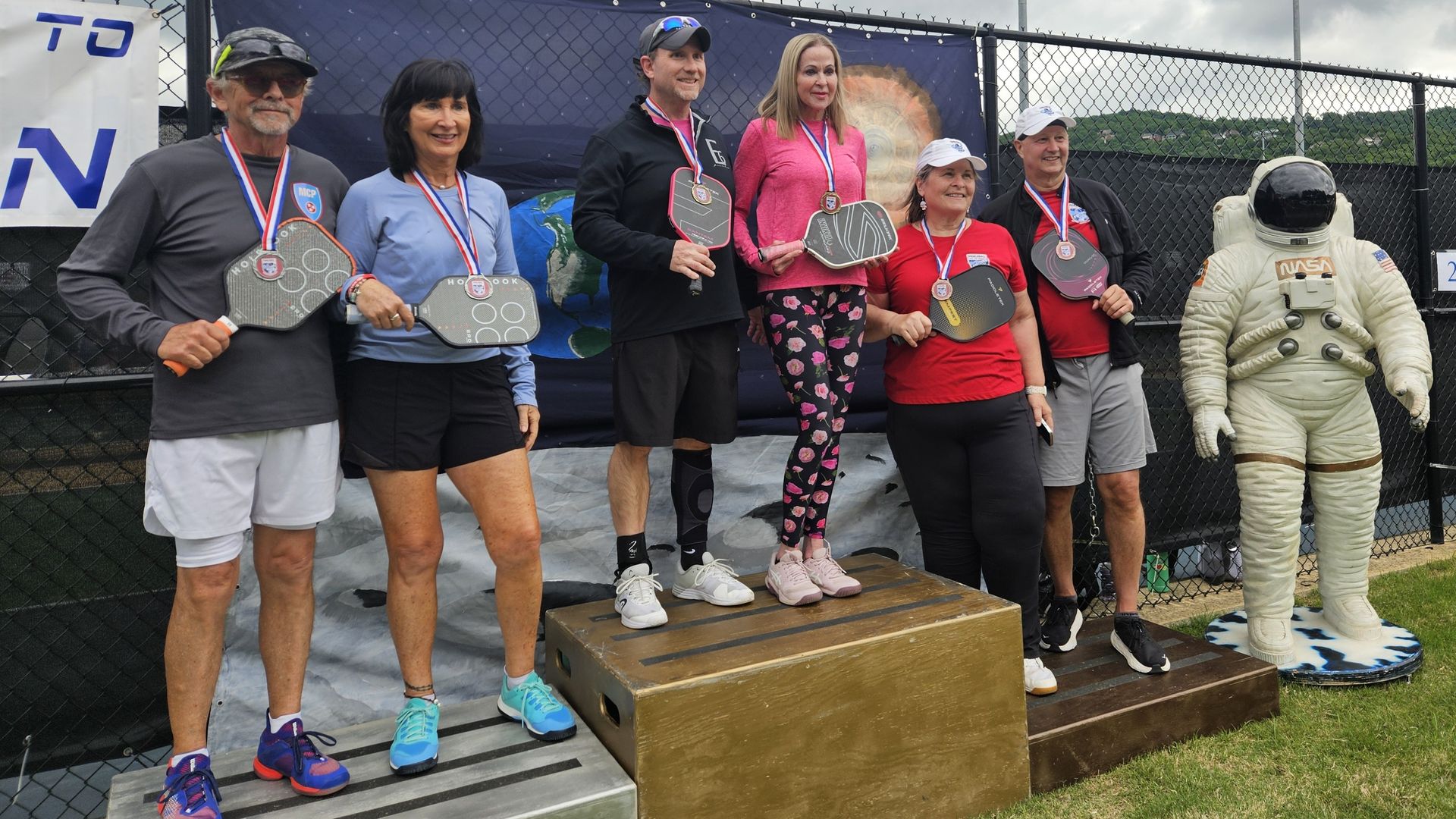 Group of athletes on an outdoor winners' podium wearing medals, holding large plaques; cloudy sky, chain-link fence with banners, and tall floodlights in the background; a person in an astronaut suit on the right.