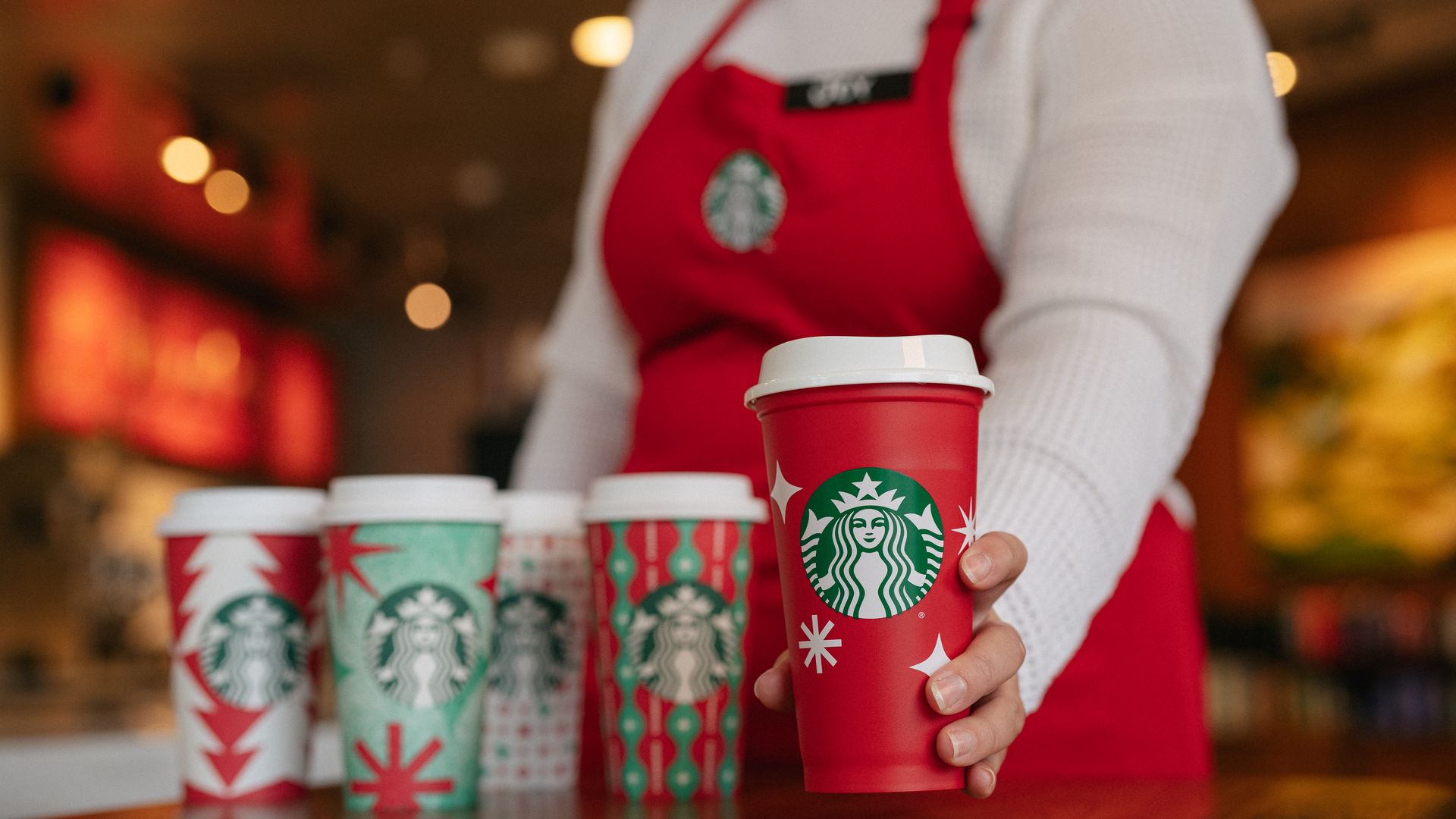 Starbucks barista holds a red reusable cup with other holiday cups nearby