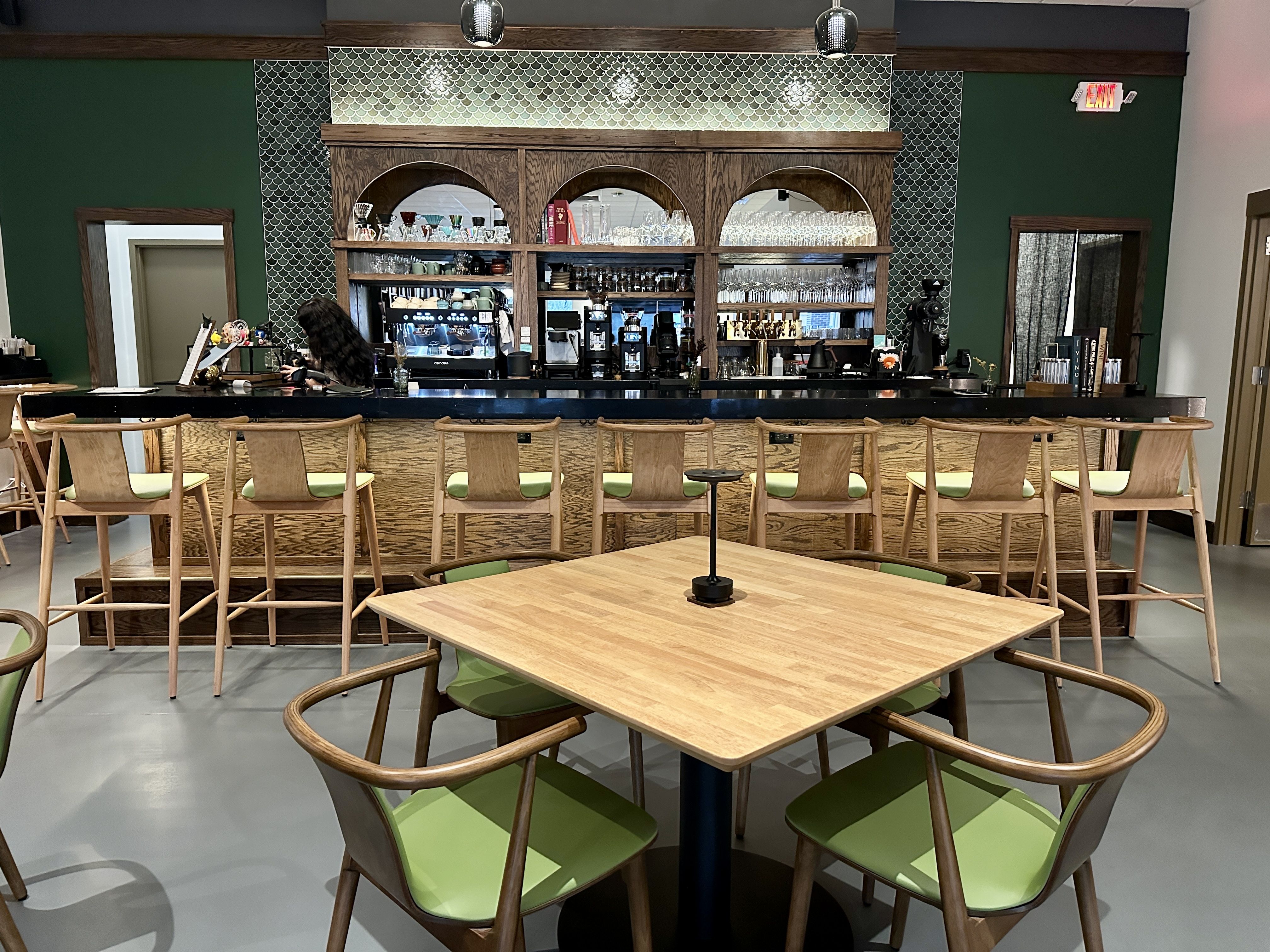 Modern coffee bar with wooden counter and green cushioned tall stools, wooden table with matching green cushioned chairs in front, dark green walls and patterned tile backsplash, person working behind bar.