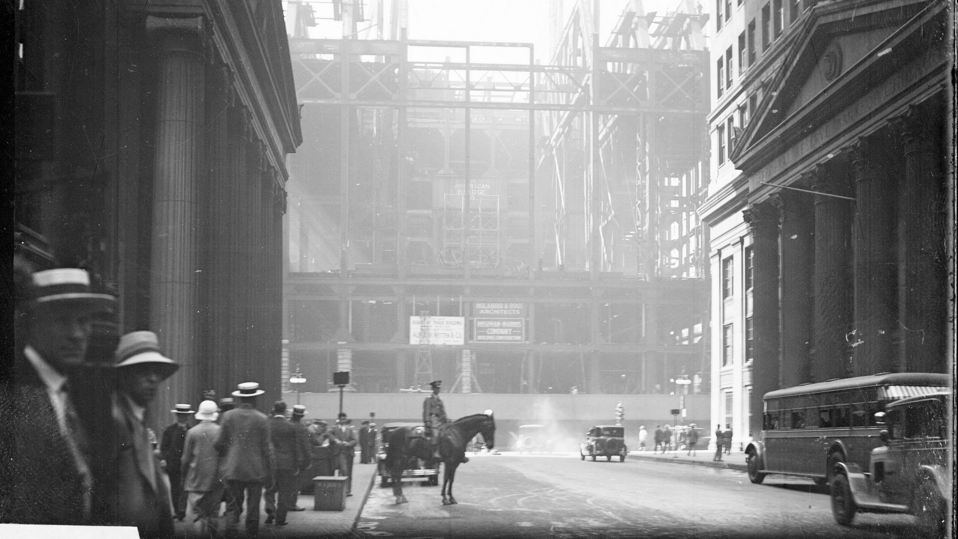 Black and white photo of a busy urban street with people in hats, a mounted police officer, early automobiles, and a large building under construction in the background.