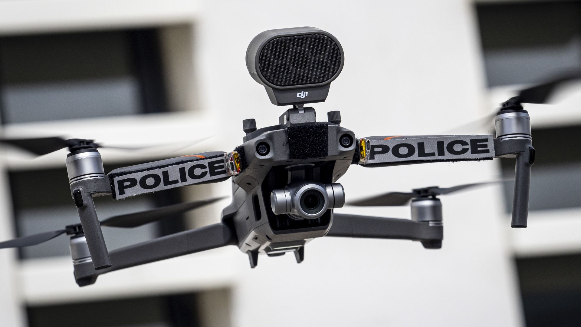 Close-up of a police drone with a large black oval speaker mounted on top, the DJI logo visible, and POLICE tape along the frame, against a blurred white building backdrop.