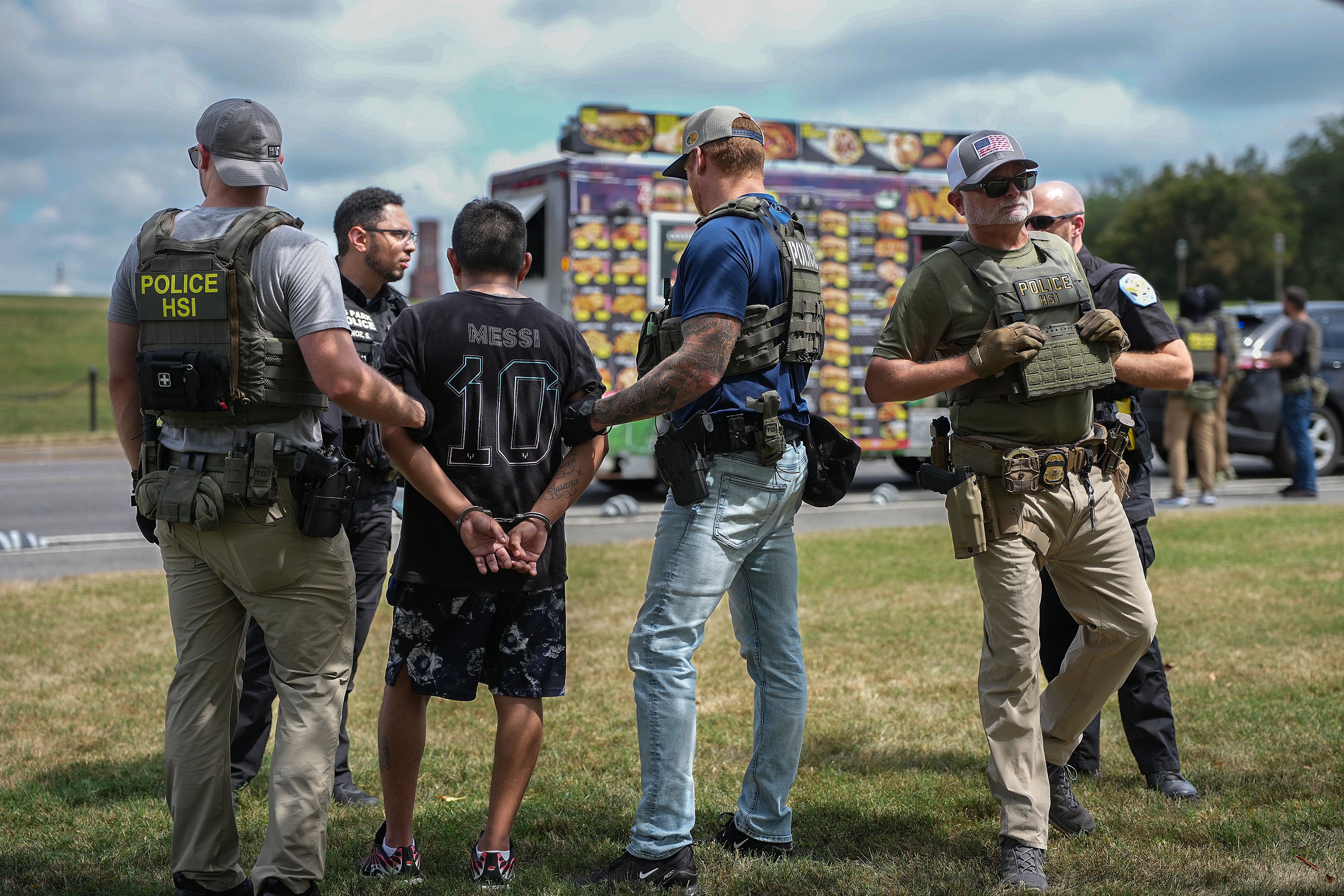 Several police officers in tactical gear arrest a man wearing a black "Messi 10" shirt and black shorts, standing on grass near a colorful food truck under a cloudy sky.