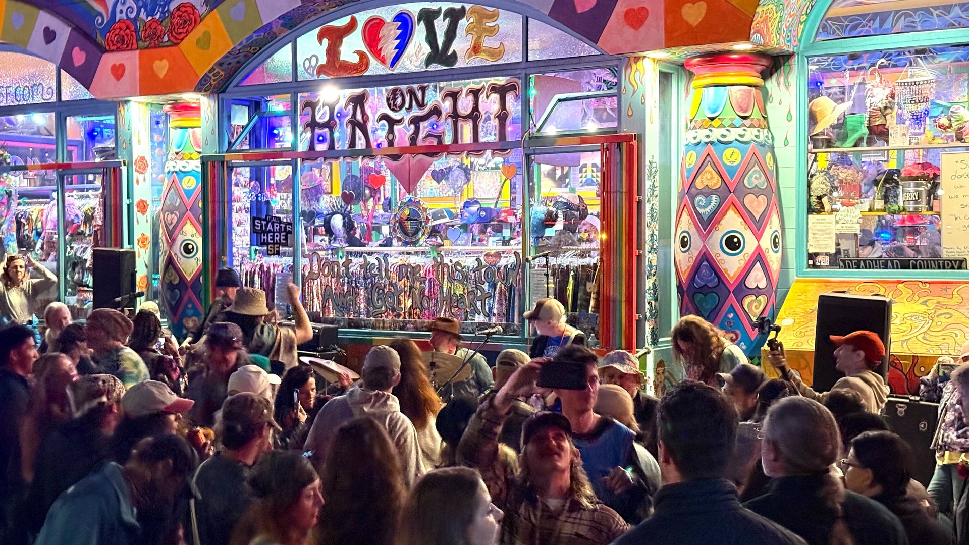 Crowd gathers outside a colorful, heart- and rainbow-decorated storefront with signs reading "LOVE ON HAIGHT" and "Don't tell me this town ain't got no heart" during a lively event at night.
