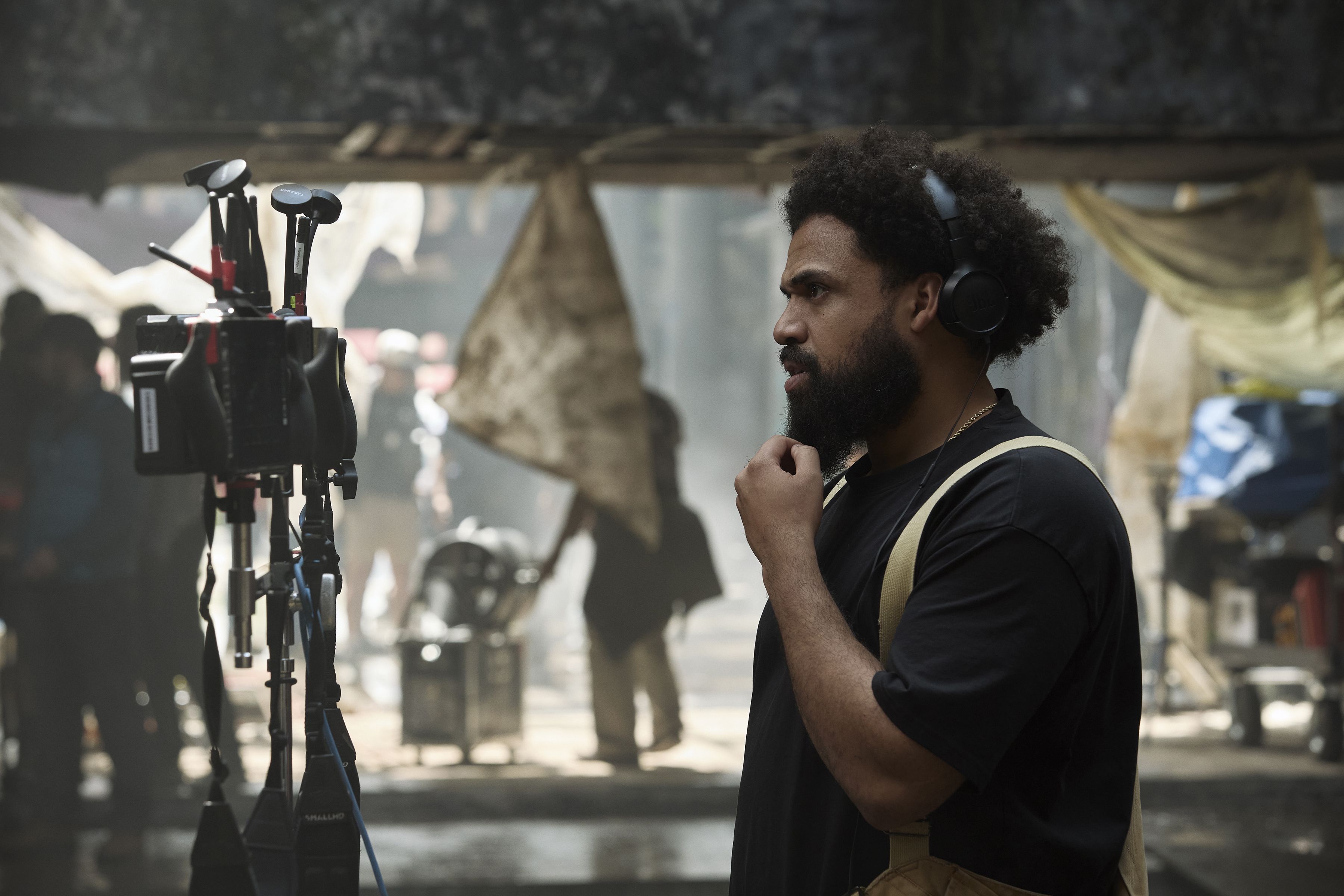 Bearded man in a black shirt with tan suspenders stands in profile, wearing headphones on a film set. Camera rigs on the left; blurred crew and a rustic backdrop.