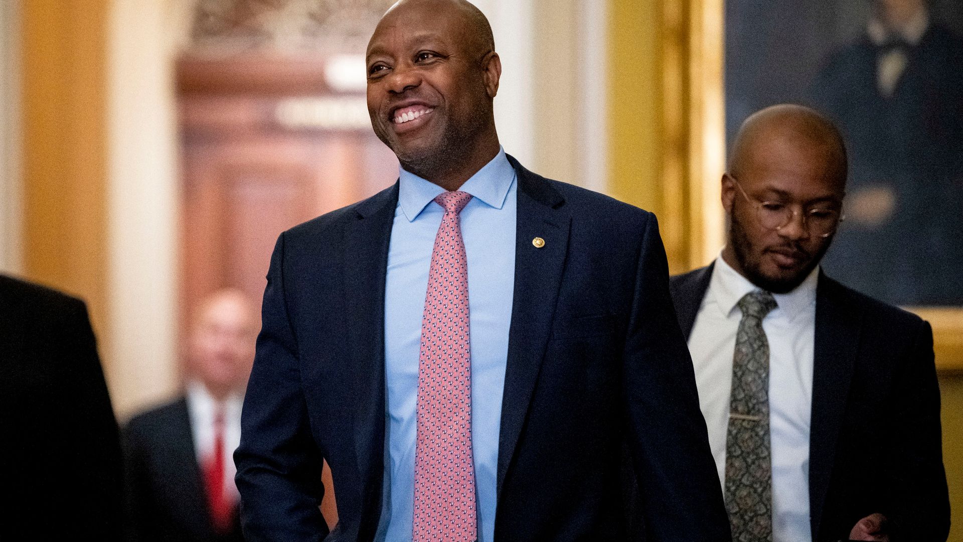 A bald man in a suit with a light blue shirt and a pink tie — in an official looking building.