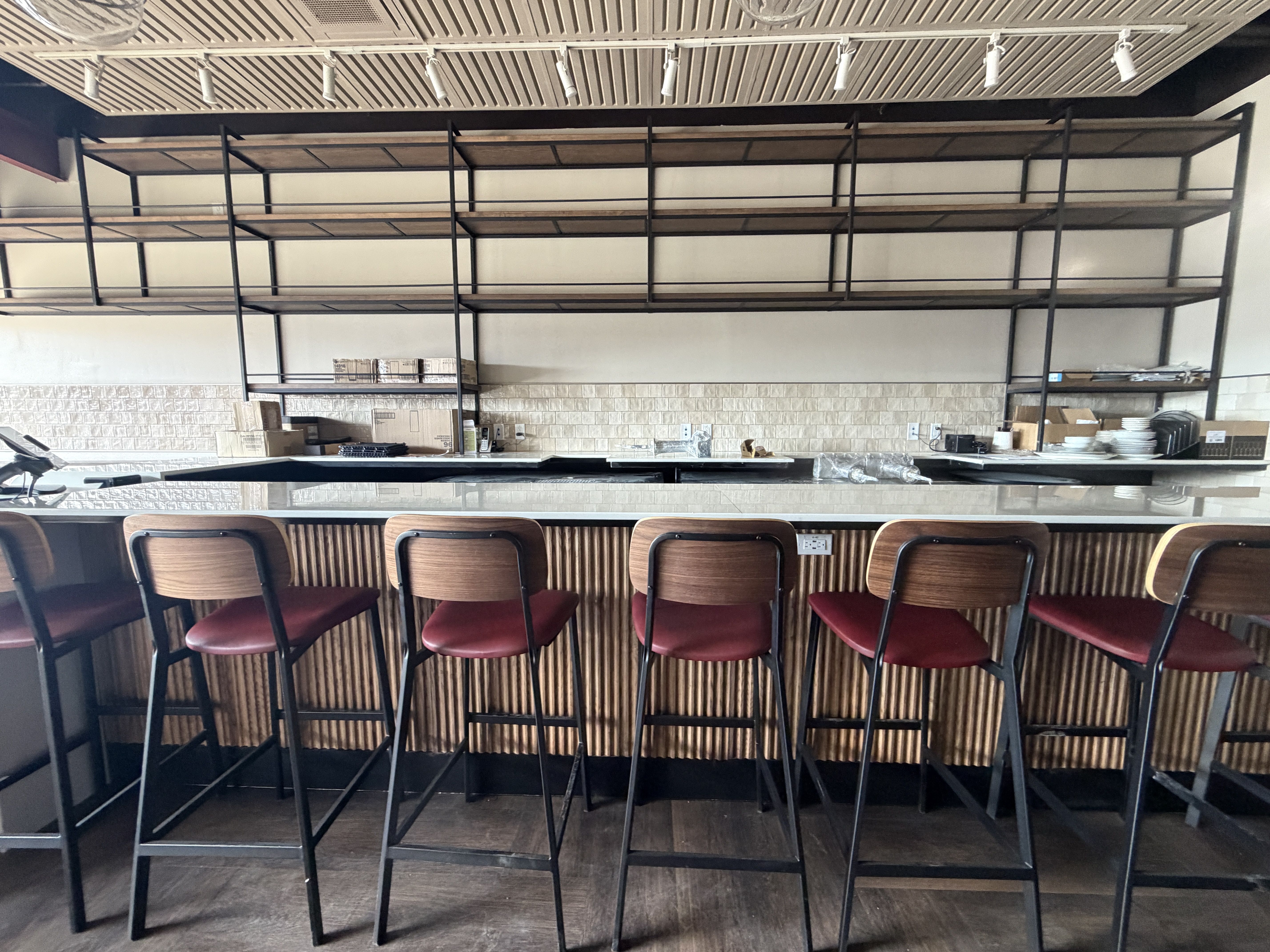 Empty modern bar area with six high stools featuring dark metal frames, wooden backs, and red seats. Behind are empty wooden shelves against a cream wall with a tiled backsplash.