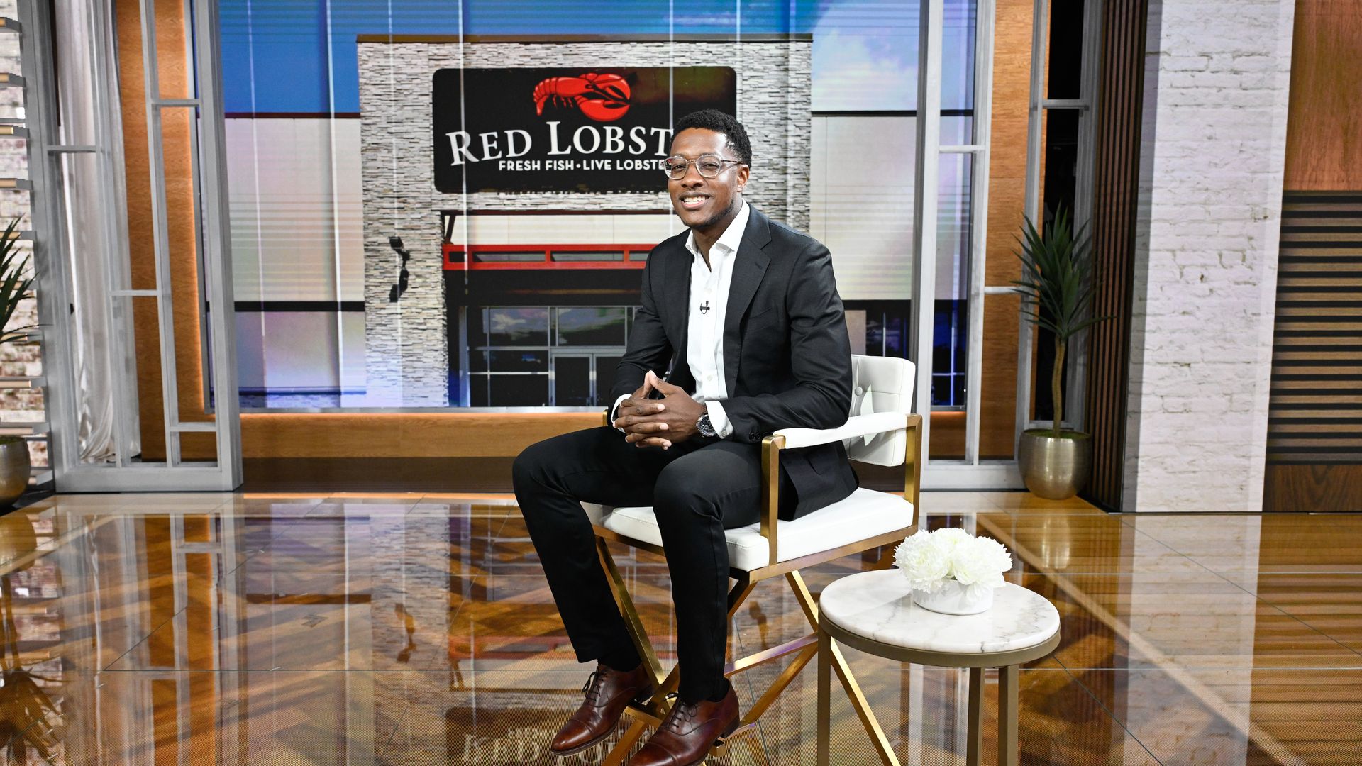 A person in a black suit and white shirt sits on a white chair in a studio; behind him a large screen shows the Red Lobster sign.