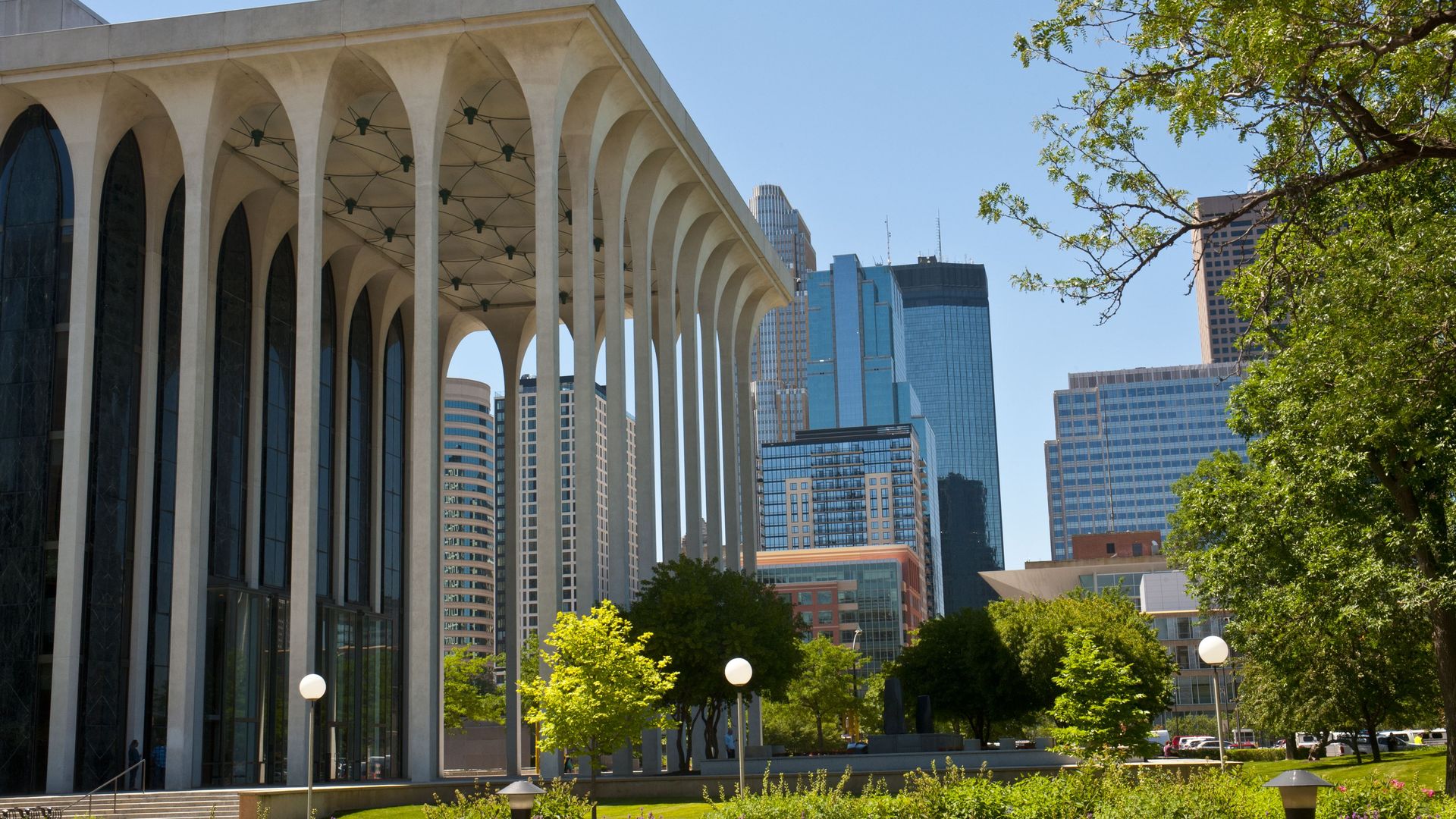 Modern urban park with green trees and flowers in foreground, tall white-columned building with arches on the left, and blue-glass skyscrapers with clear sky in background.
