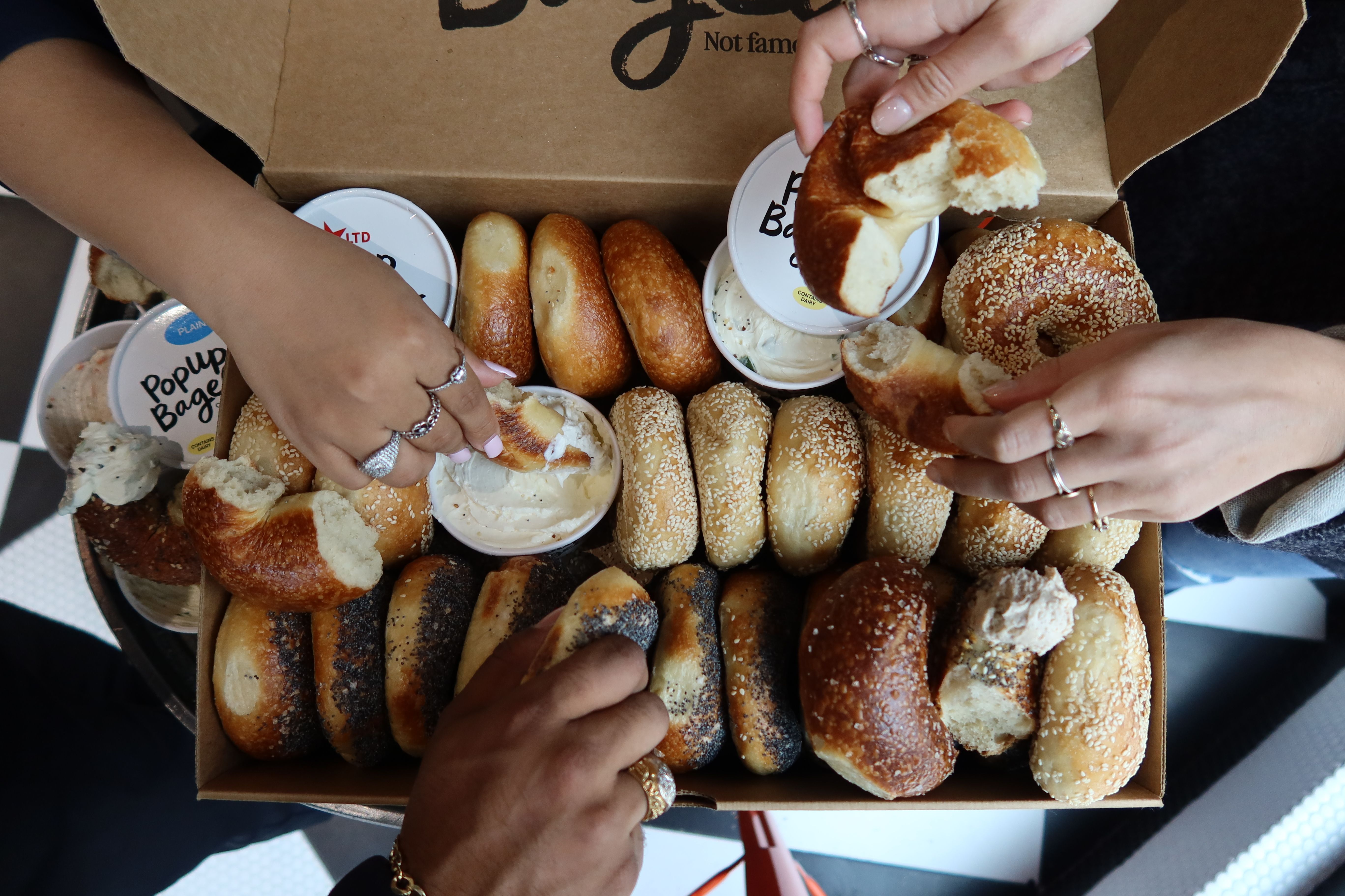 Top-down view of a box of assorted bagels (sesame, plain, poppy seed) with cream cheese cups; several hands wearing rings reach in to grab bagels.