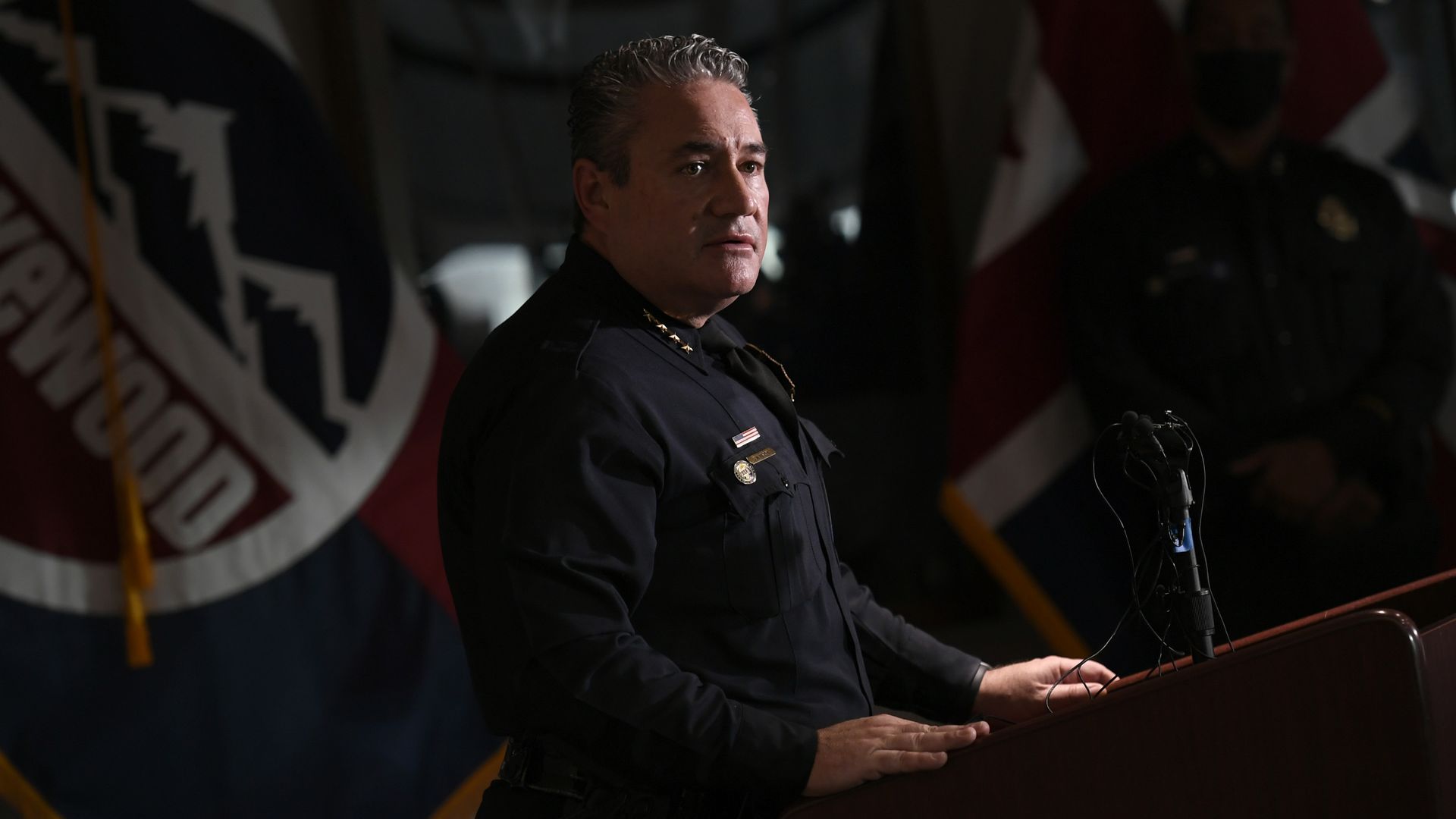 A police chief in full uniform stands near a lectern in a dimly-lit room while addressing member of the press.