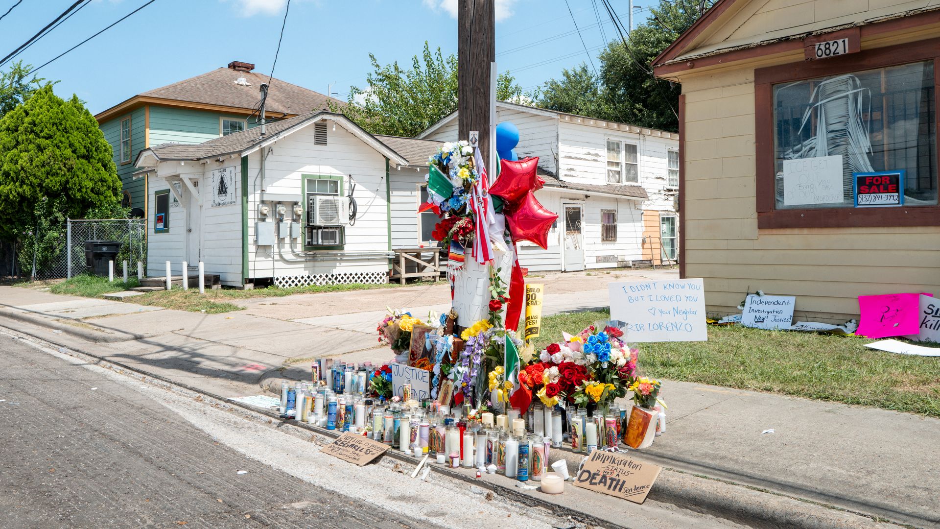 Sunny street memorial with candles, flowers, balloons, and signs at a corner; beige storefront, white houses, and overhead power lines.