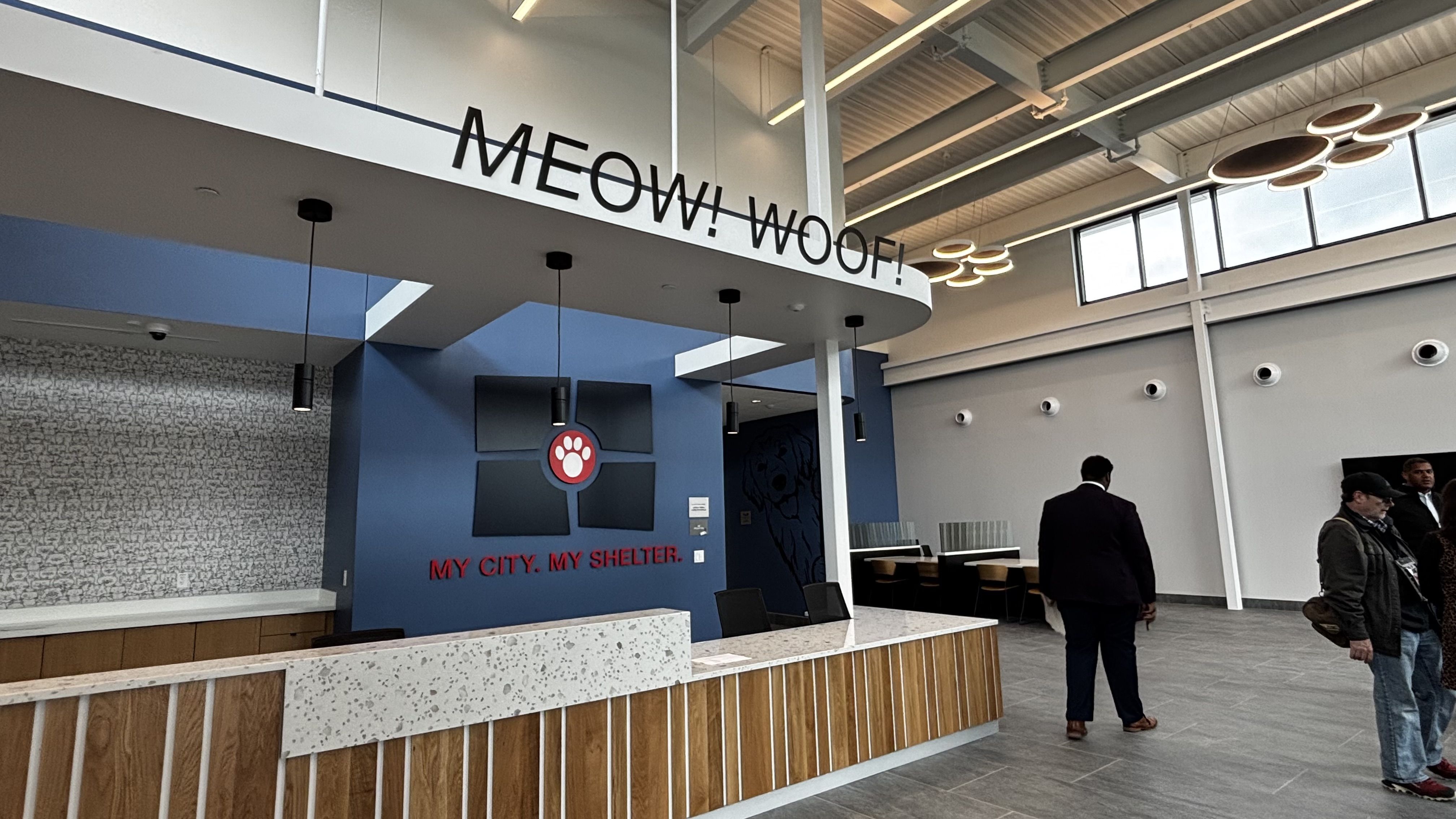 Interior of an animal shelter lobby with a curved white beam that reads "MEOW! WOOF!", blue wall with paw logo and "MY CITY. MY SHELTER.", terrazzo counter, and people nearby.