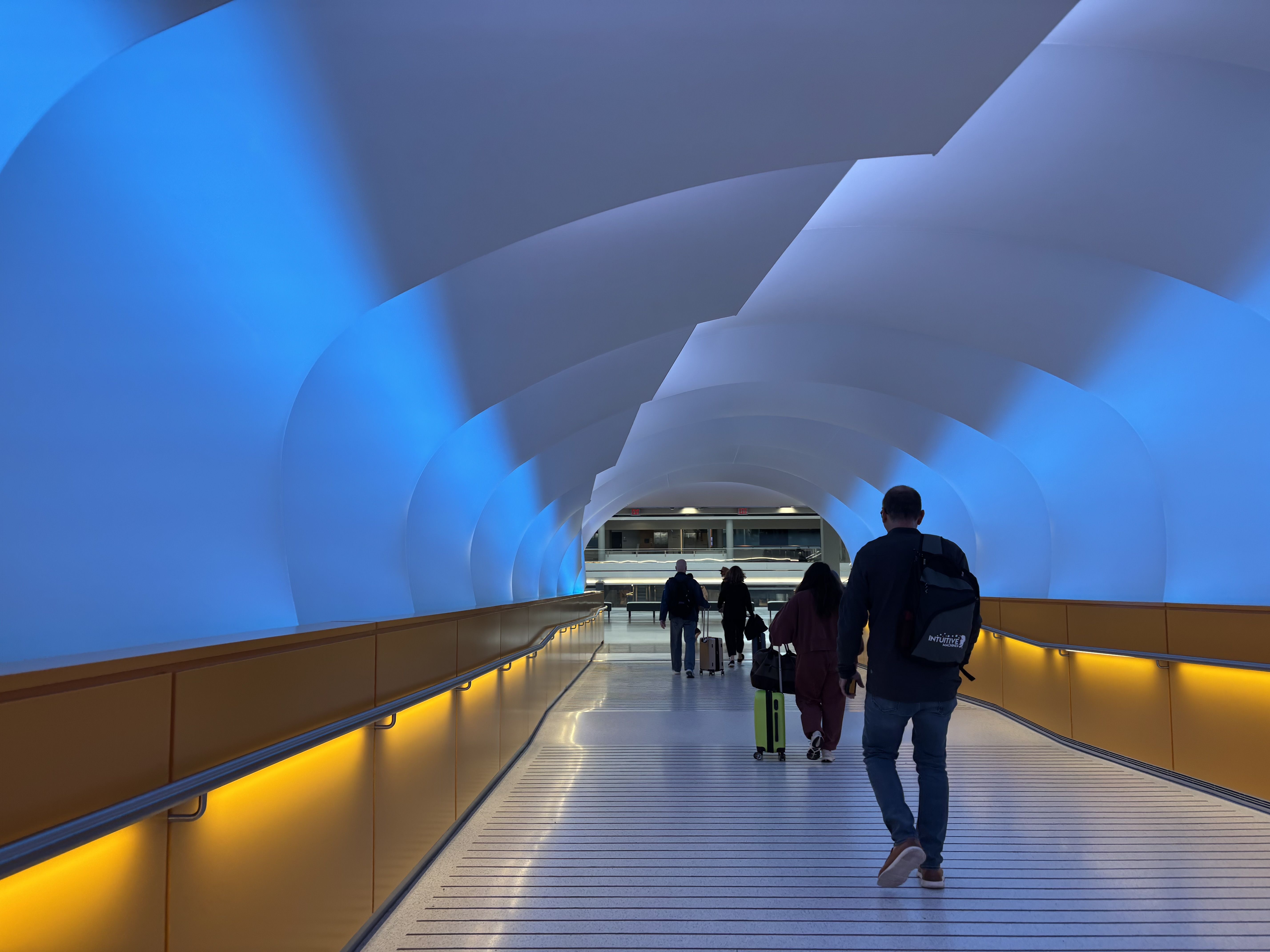 Blue-lit tunnel walkway with white curved arches, yellow side rails, and travelers pulling luggage toward the far end of a bright terminal corridor.
