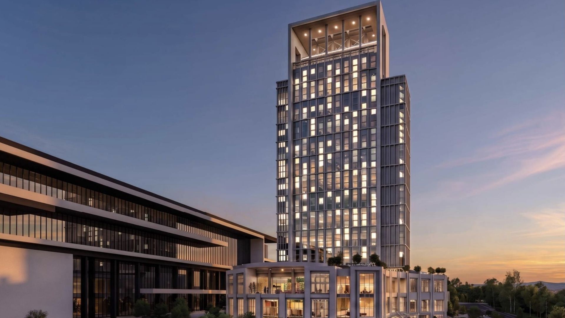 Evening view of a tall glass office tower with a rectangular framed top, its windows lit; a low winged building to the left, sunset sky, trees, and people near the entrance.