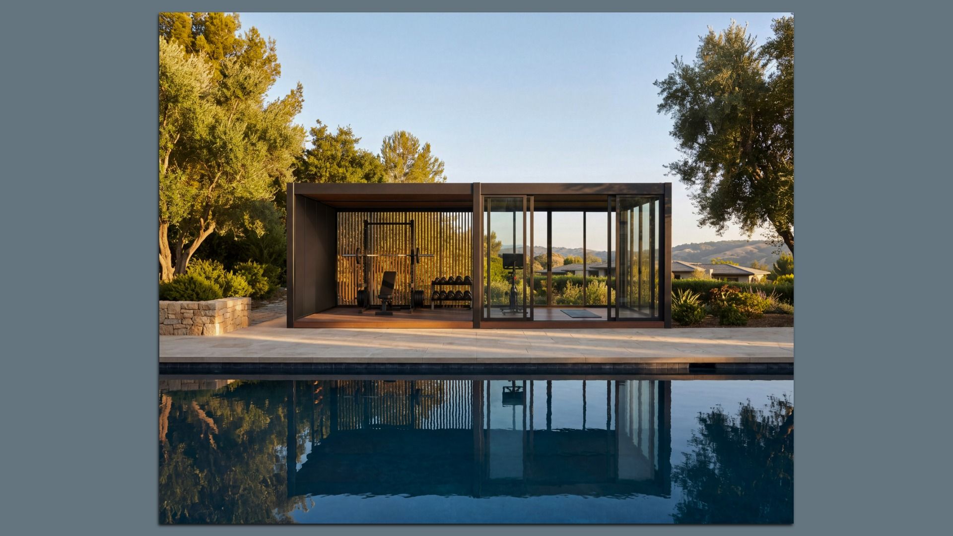Open-air gym pavilion with a dark metal frame and glass walls beside a pool. Inside, weights, a bench, and a barbell rack are visible; trees and distant hills glow in warm evening light.