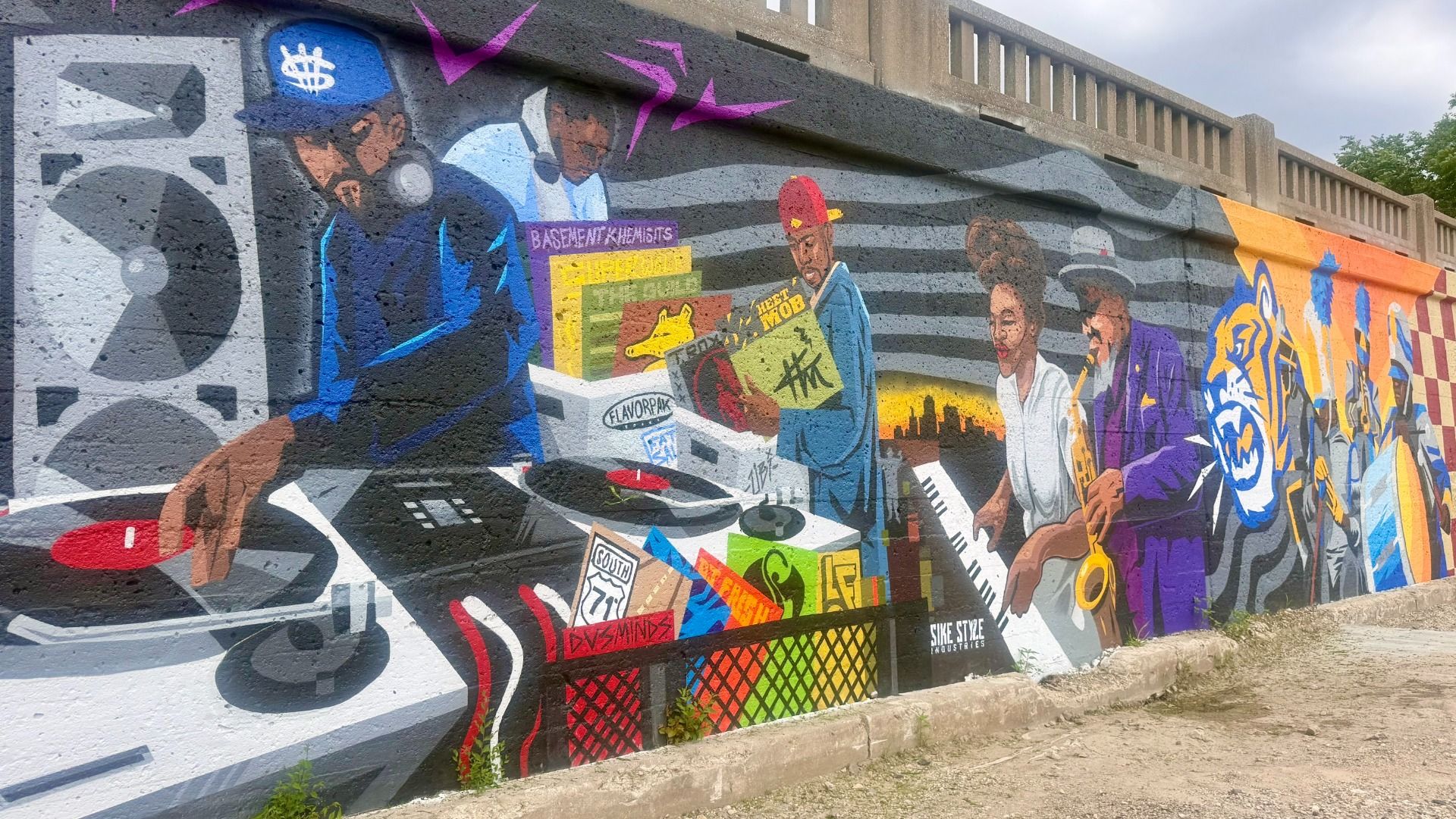 Vibrant mural on a concrete wall shows a DJ at a turntable, a singer with a microphone, a keyboard player, and a tiger mascot among graffiti shapes in blue, purple, orange, and yellow.