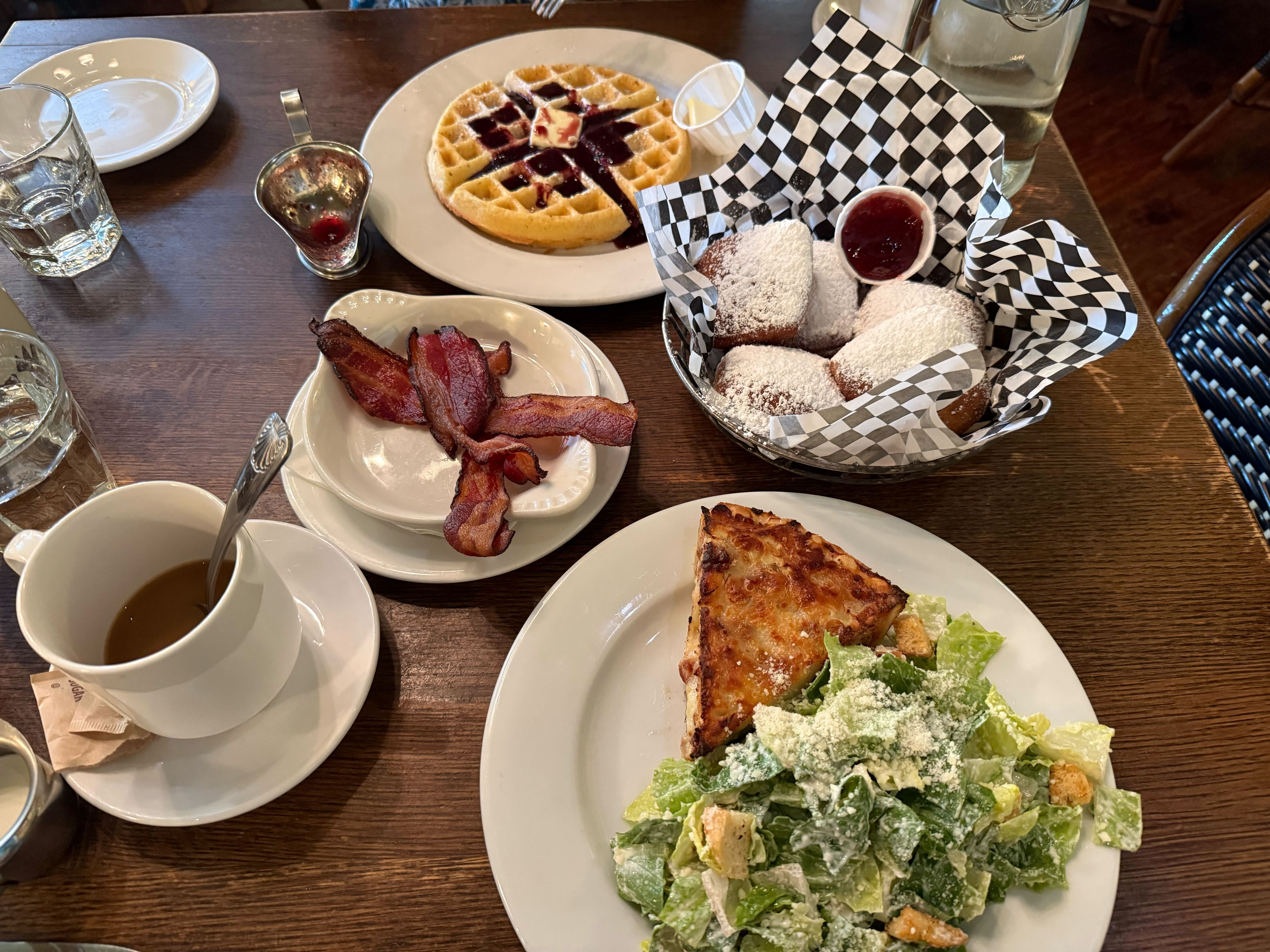 Top-down view of a breakfast spread: a waffle with berry sauce, powdered donuts in a black-and-white checkered basket, crispy bacon on a plate, a quiche slice with a side salad, and coffee.