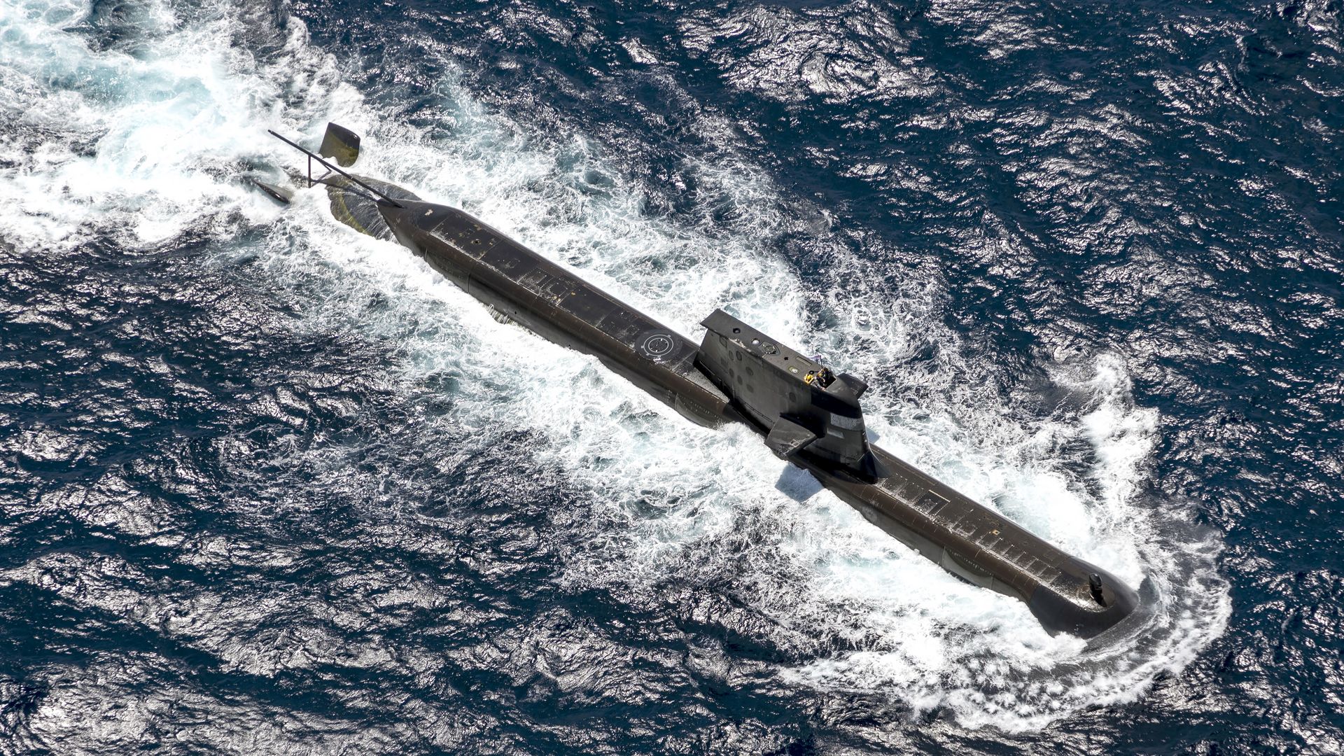 A black submarine cruising on the ocean surface, creating white foamy waves as it moves through deep blue water under sunlight.