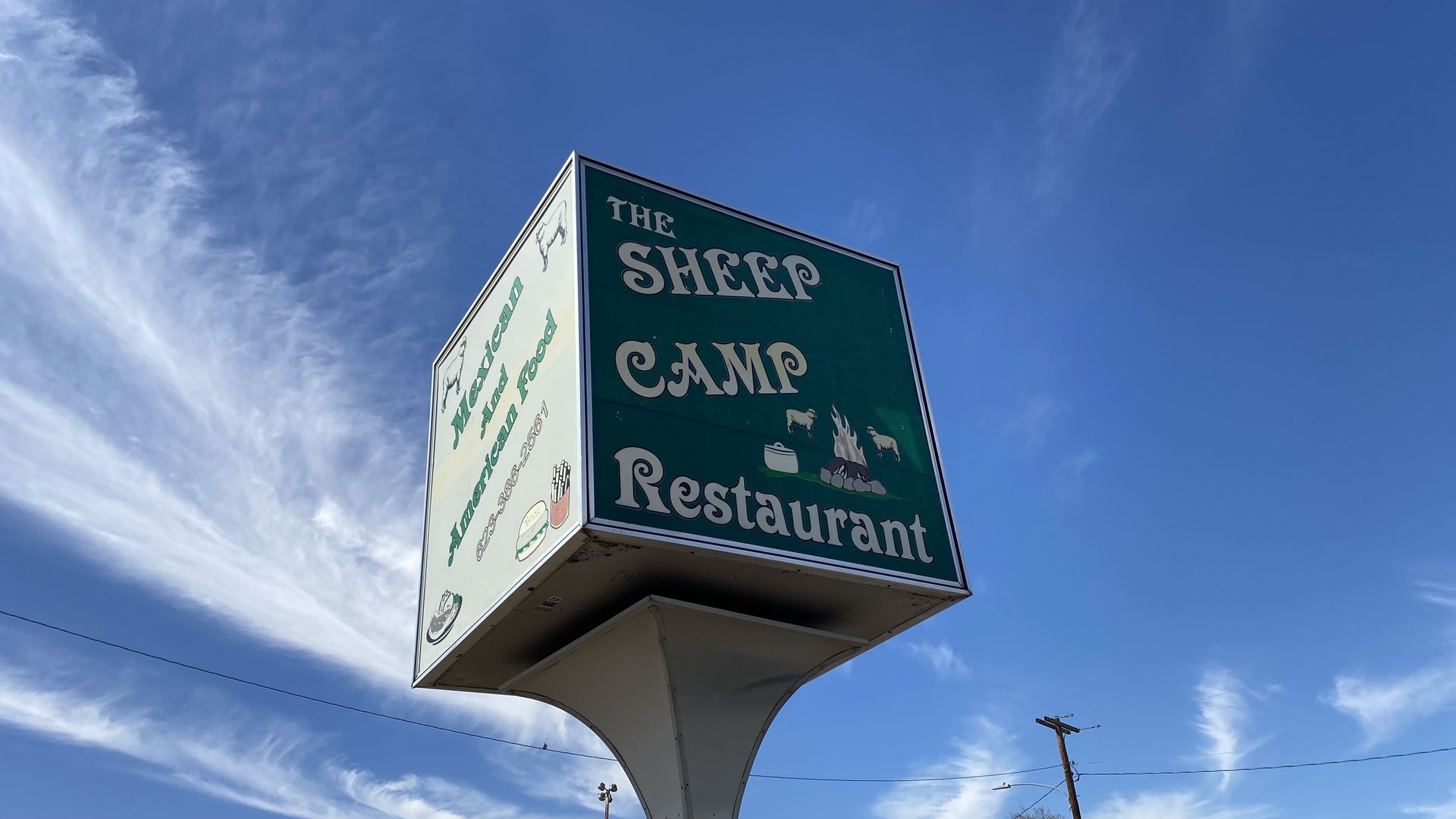 A green, cube-shaped sign atop a pole advertising a restaurant called The Sheep Camp. 
