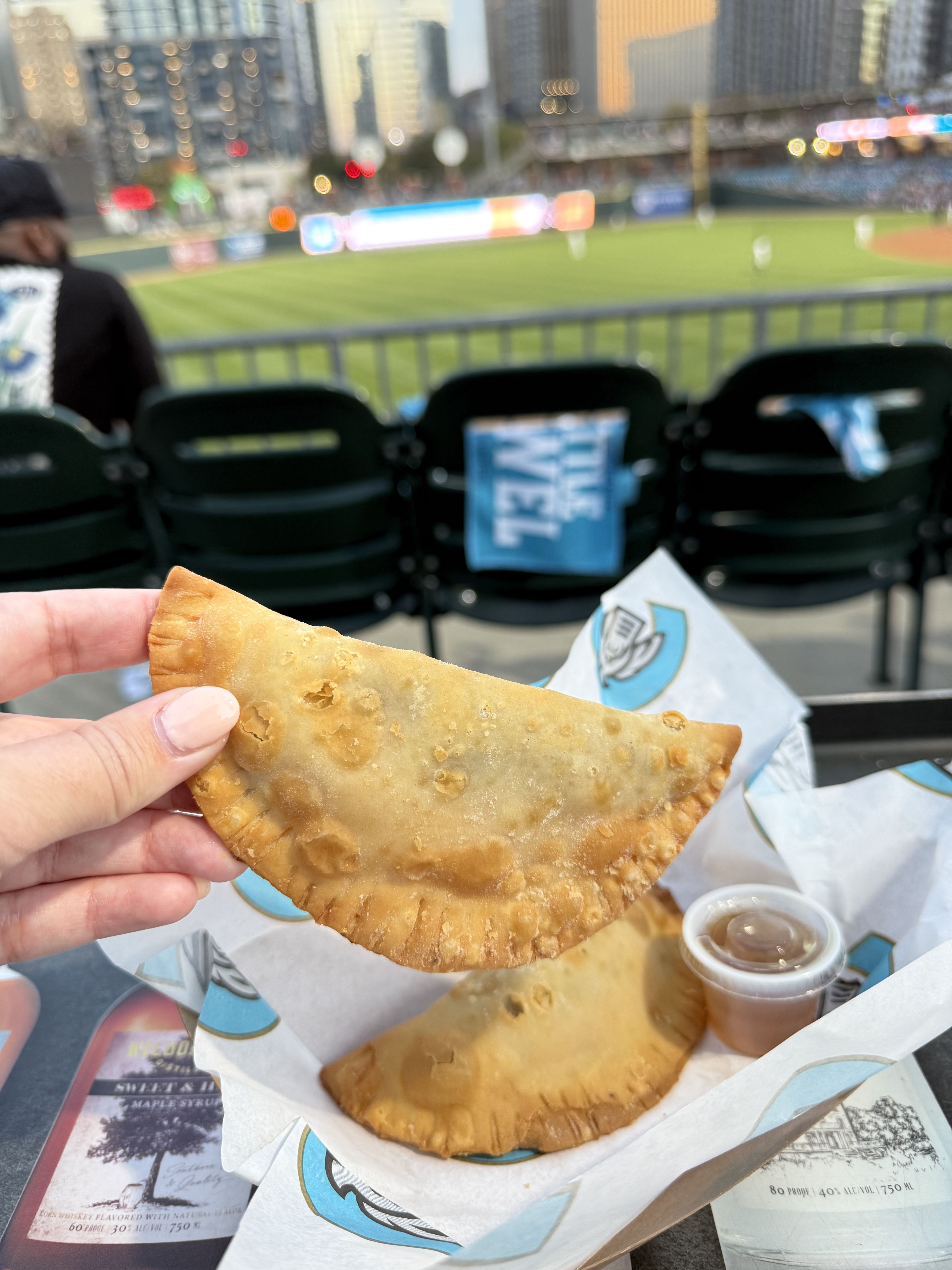 A person holds a golden-brown empanada above a tray with another empanada and dipping sauce at a baseball game.