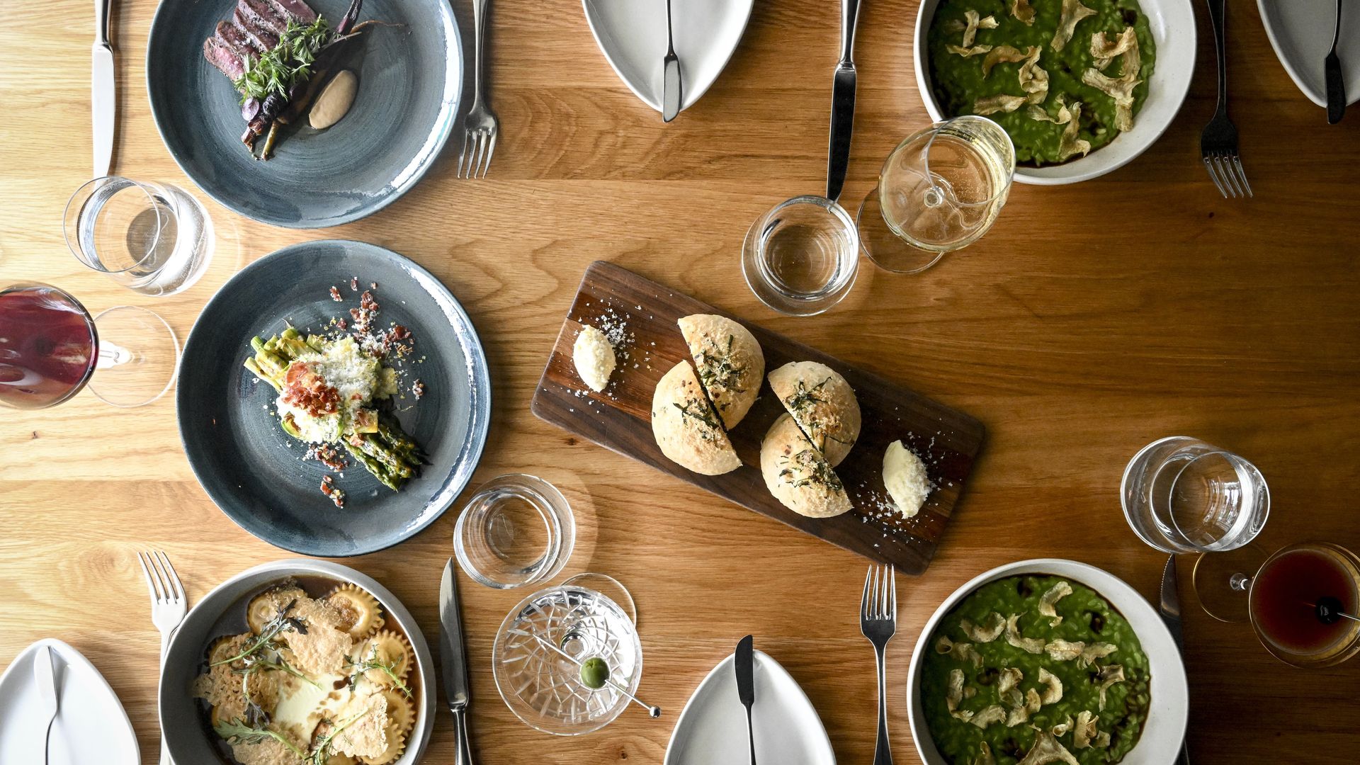 Top-down view of a wooden dining table set with blue-gray plates of sliced meat, bowls of green soup, a bread board with rolls, and scattered wine glasses, forks, and knives.