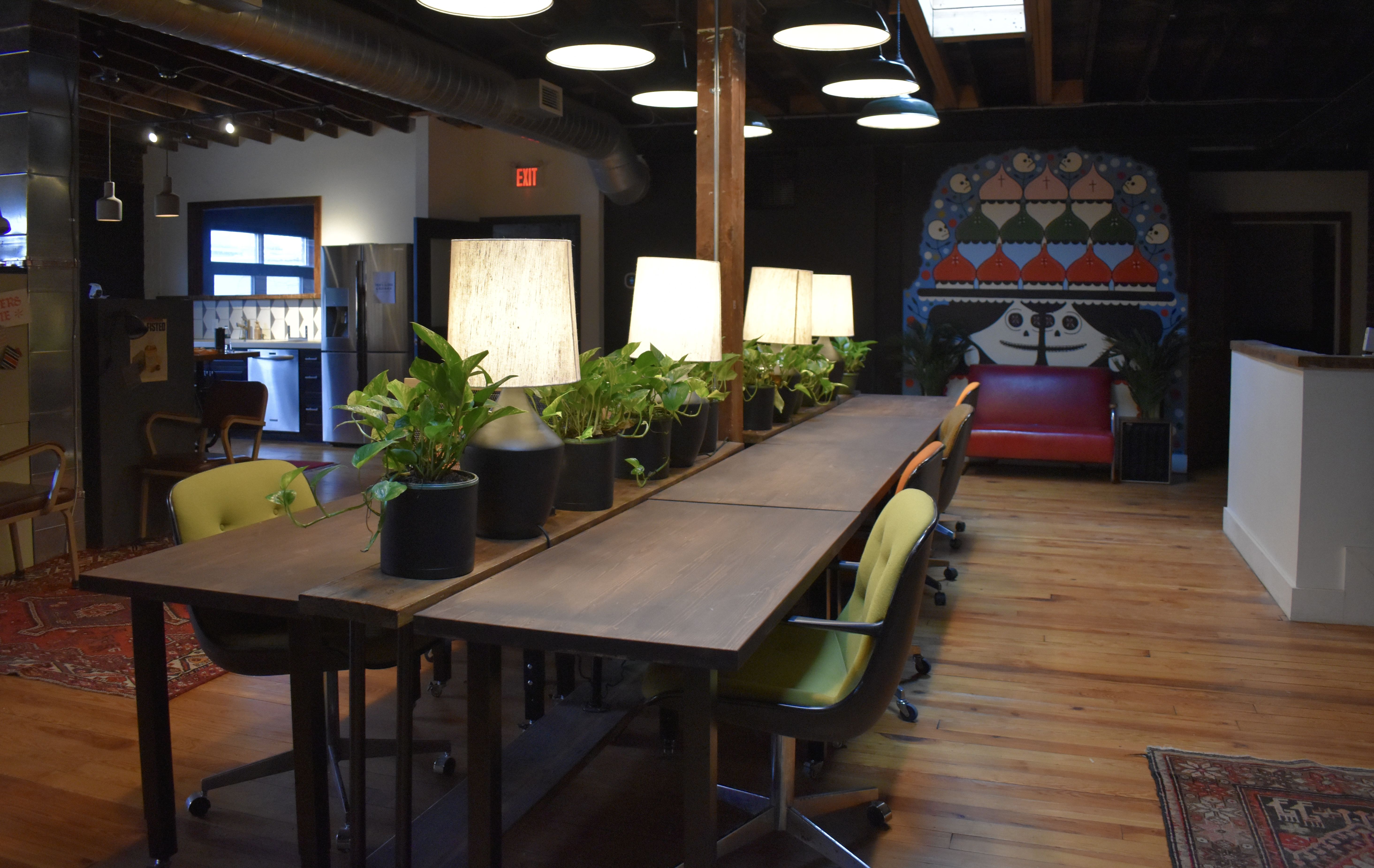 Cozy office space with wooden floors, a long wooden table with green swivel chairs, potted plants, lit lamps, and colorful wall art behind a red couch in the background.