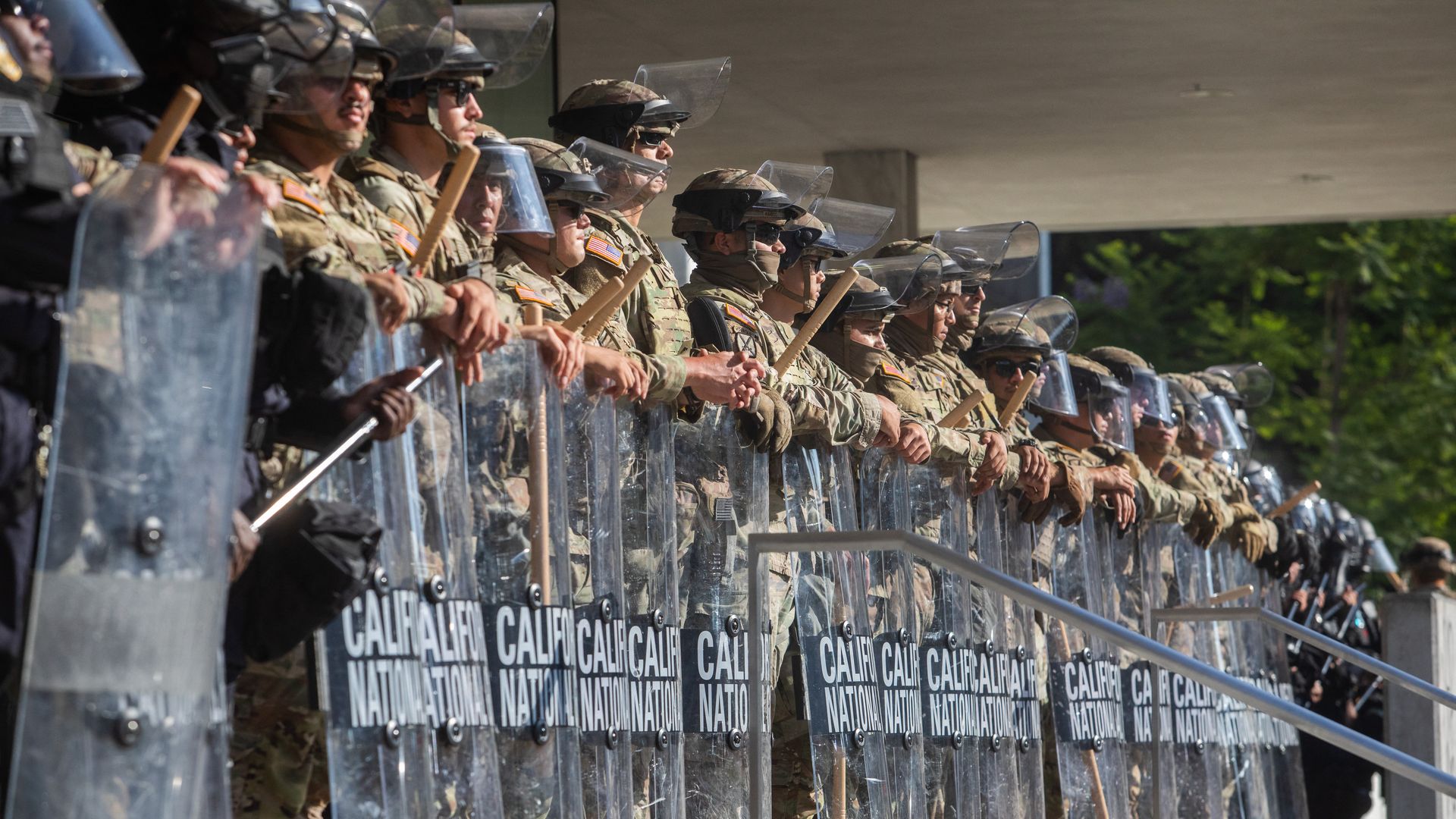 Membri della Guardia Nazionale della California sono in fila davanti a un edificio federale, armati di scudi antisommossa.