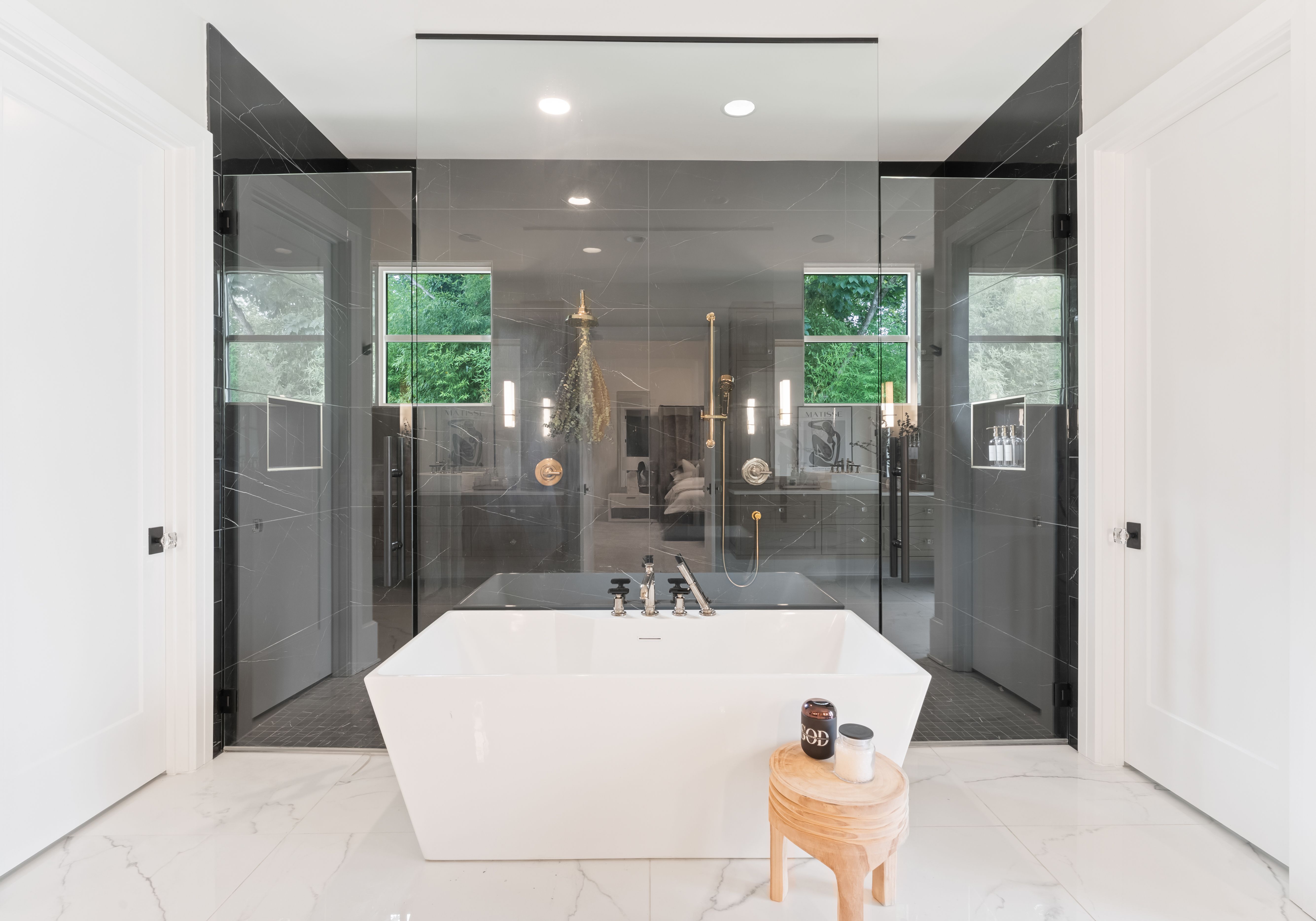 Modern bathroom with white freestanding tub, wooden stool with bath salts, large glass shower with black marble walls, two windows showing green trees, and white marble floor.