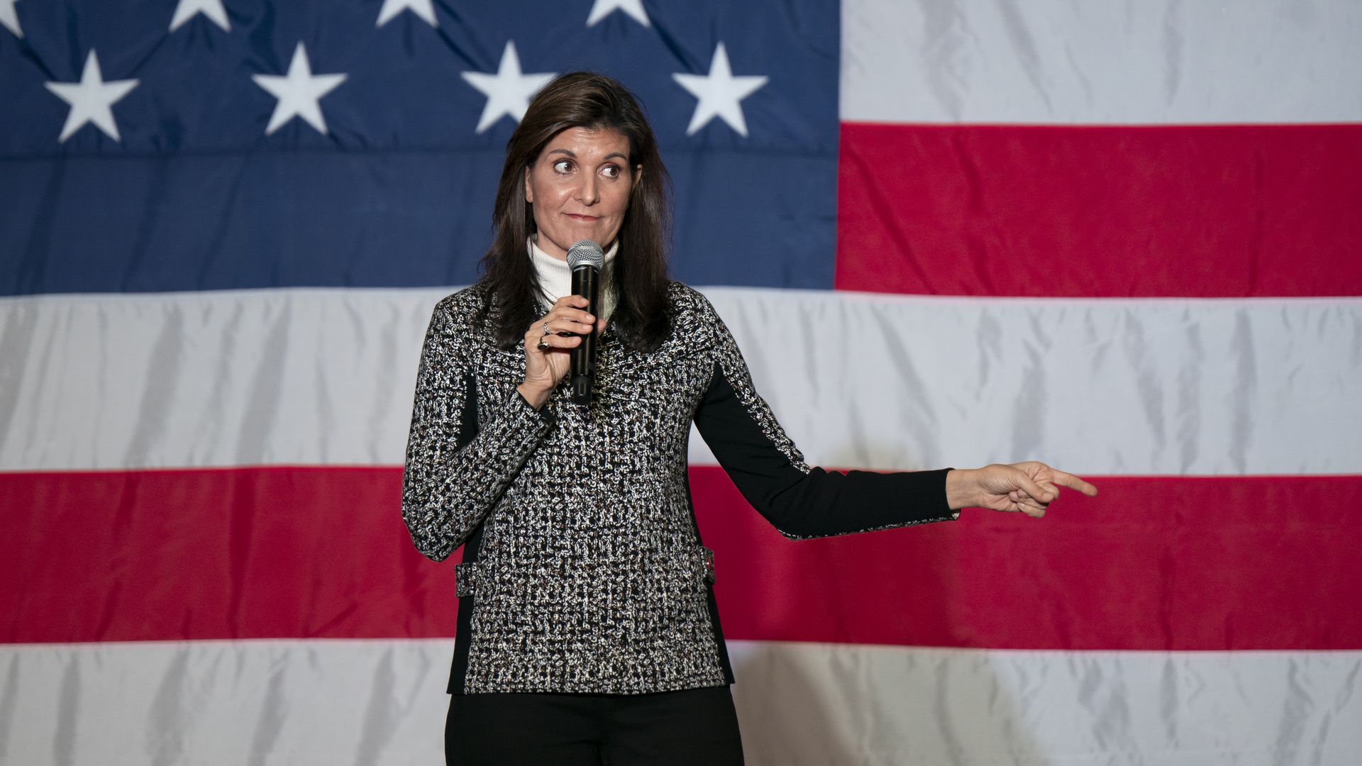 A woman holds a microphone while standing in front of a large American flag backdrop.