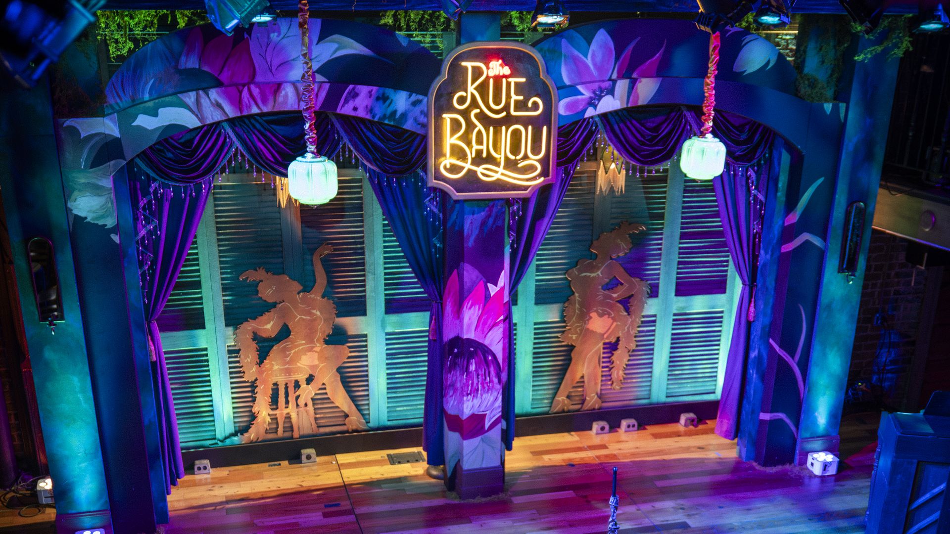 Stage lit in blue and purple with teal accents; neon "Rue Bayou" sign above purple curtains, orange dancer silhouettes on back wall, wooden floor, and a circular platform with a microphone stand.