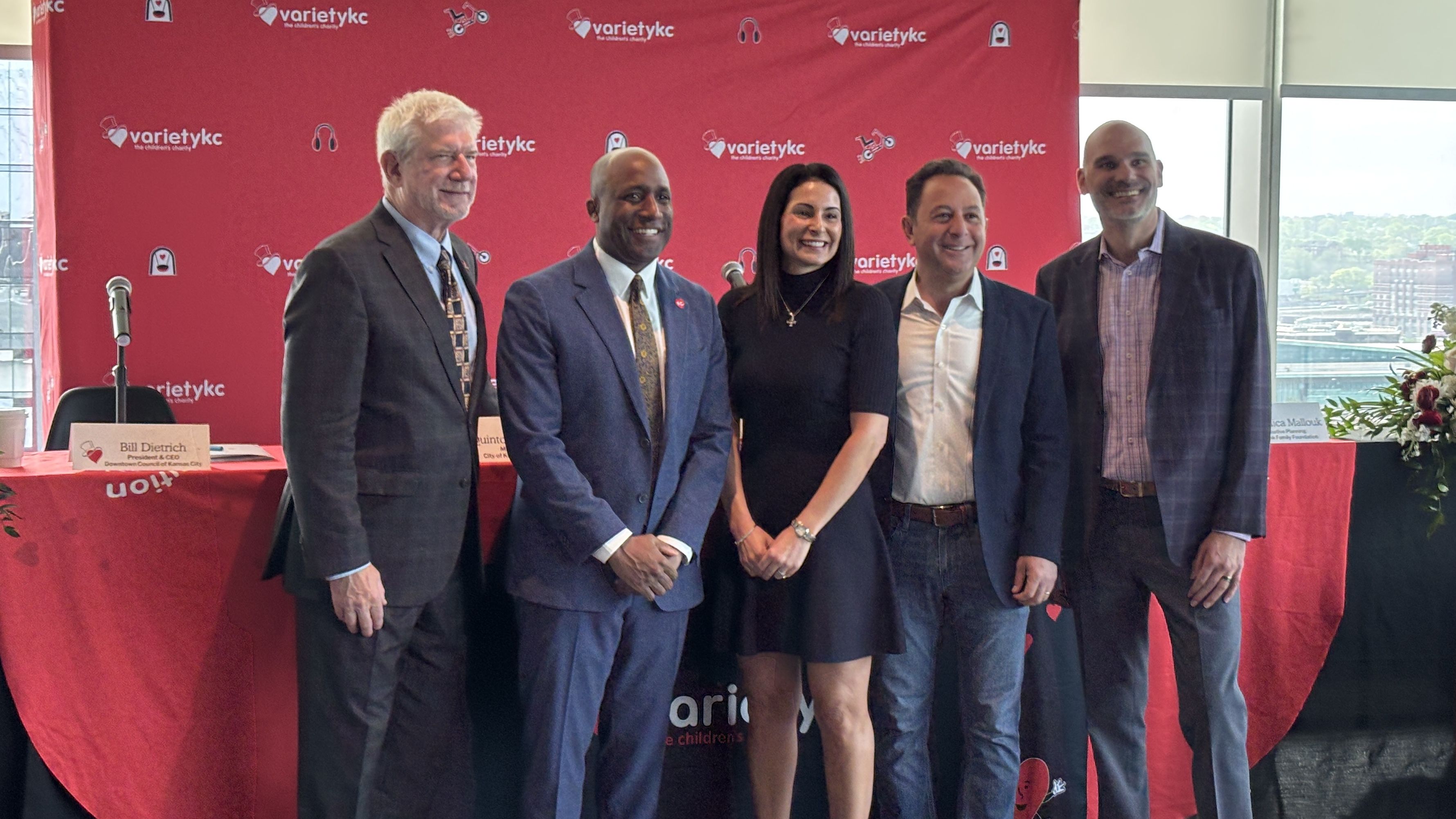 Five professionally dressed individuals pose in front of a red VarietyKC backdrop at a panel; a microphone on a stand sits near a banner-covered table and a cityscape is visible through the windows.