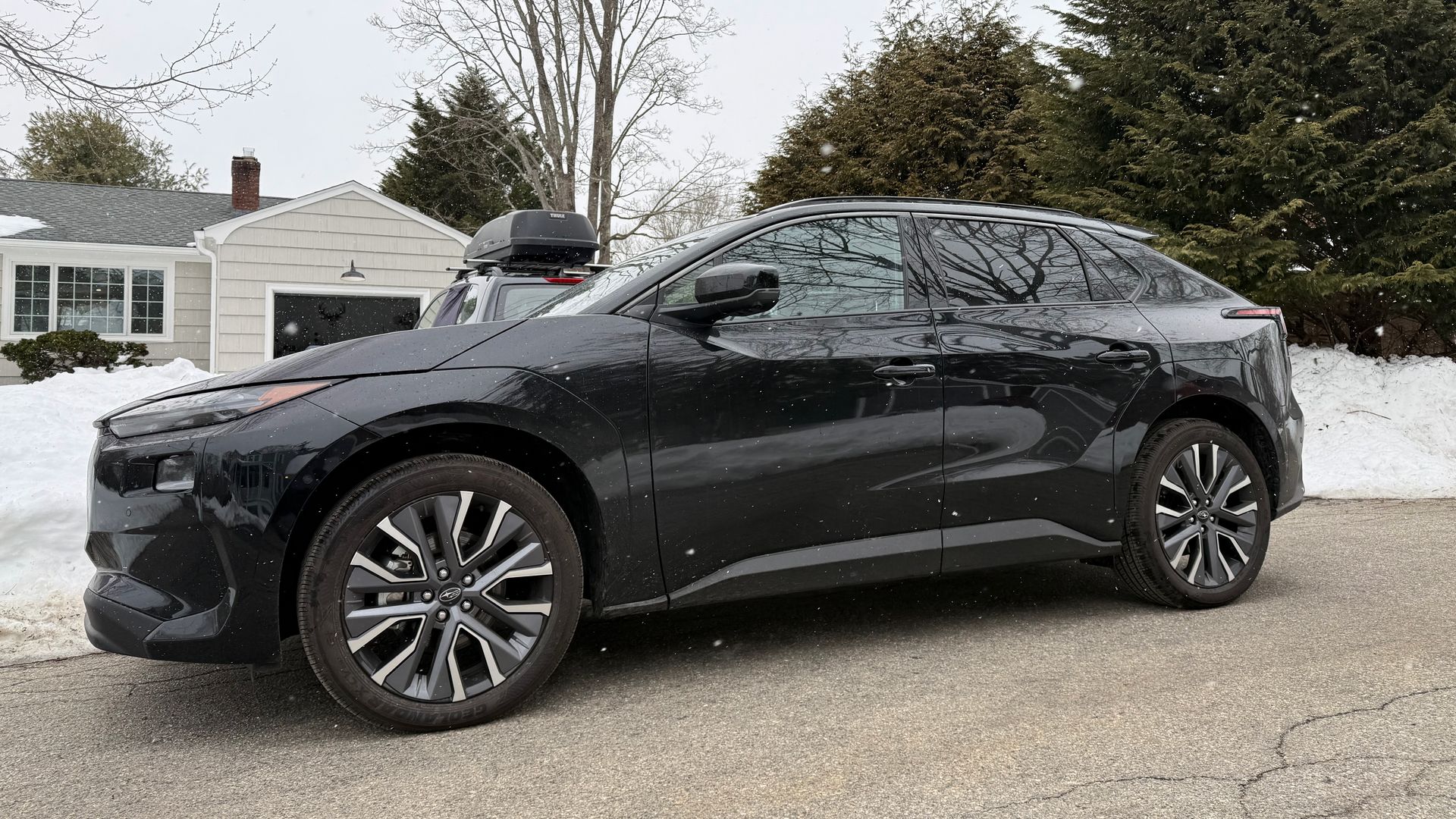Black Subaru Solterra electric SUV parked on a snowy suburban street. Snow piles and bare trees line the yard, with a beige house and garage in the background.