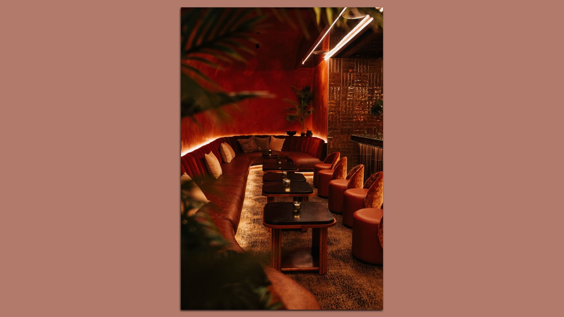 Dim lounge with a long red curved sofa along the left, small black tables with candles, and brown stools on the right. Warm lighting, plants, and a metallic wall create an intimate bar vibe.